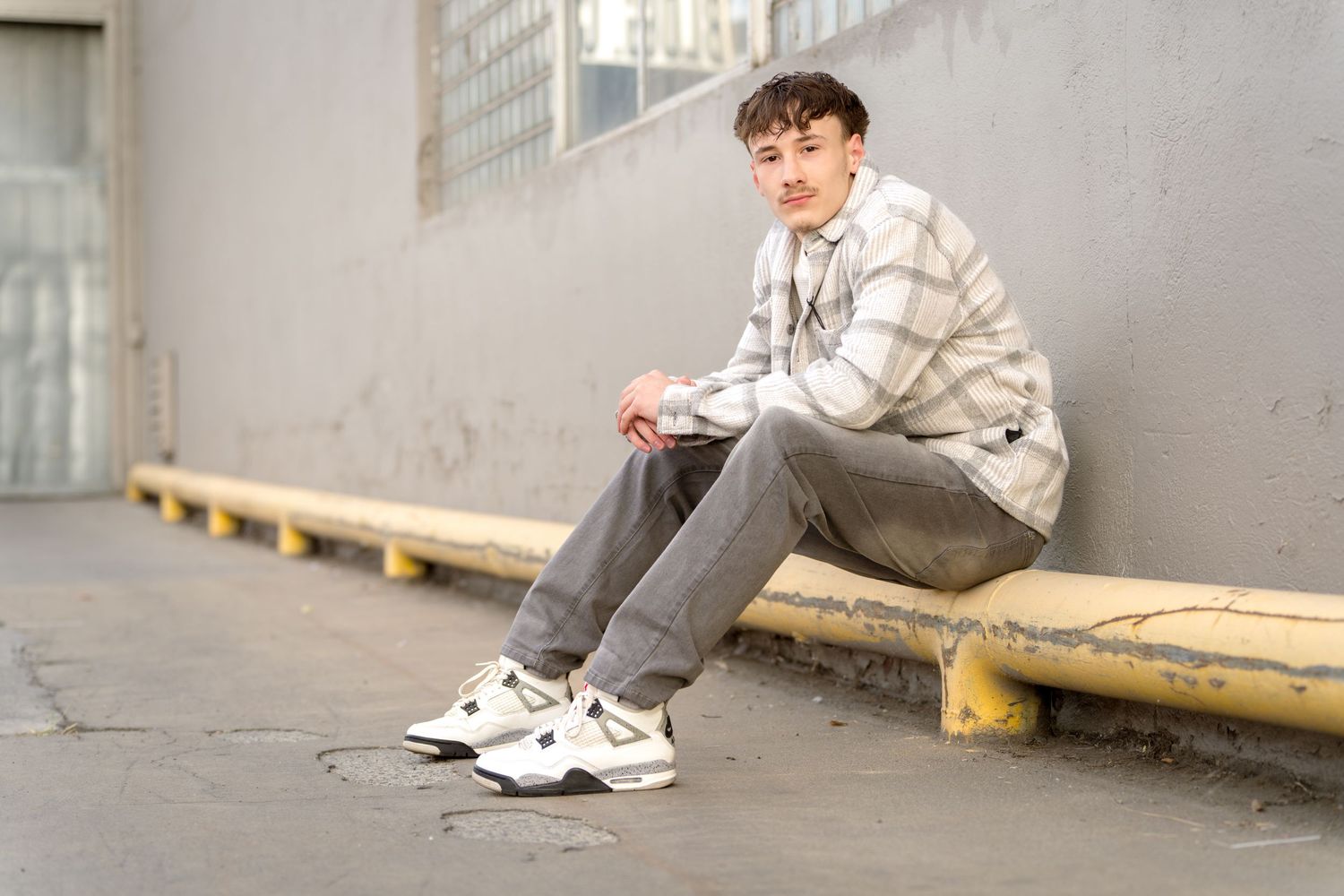 A senior guy sits on the ground against a gray wall.
