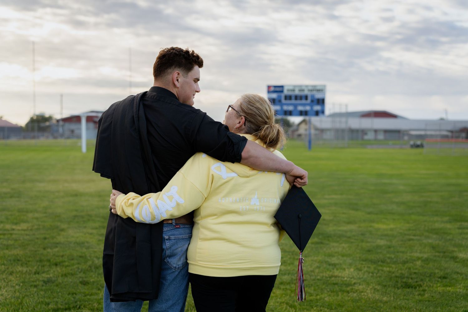 A graduate in cap and gown embraces a woman in a yellow shirt on a green field at dusk.