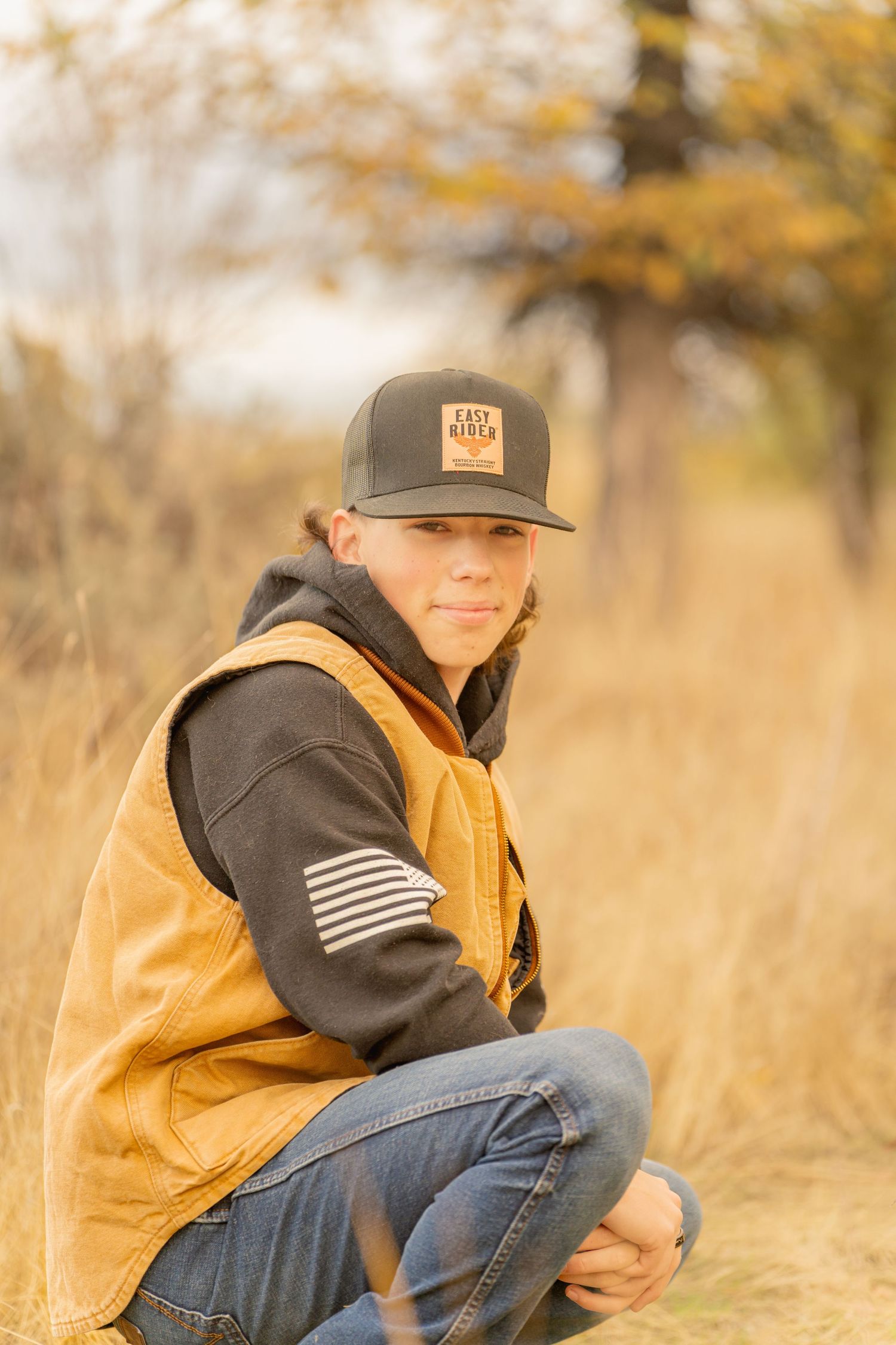 Person wearing a brown vest and baseball cap kneels in tall autumn grass with blurred golden foliage in background.