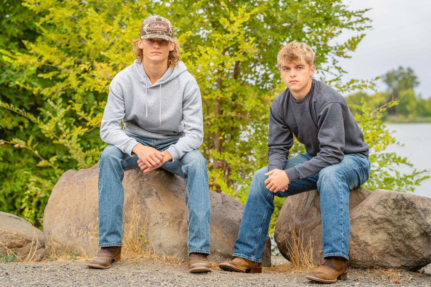 senior brothers in casual attire sit on large rocks outdoors against a backdrop of green foliage and water.