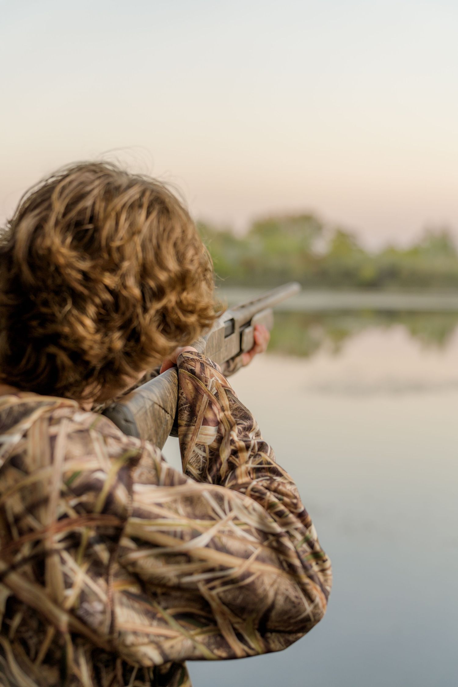 A hunter in camouflage gear aims their gun while duck hunting at sunrise.