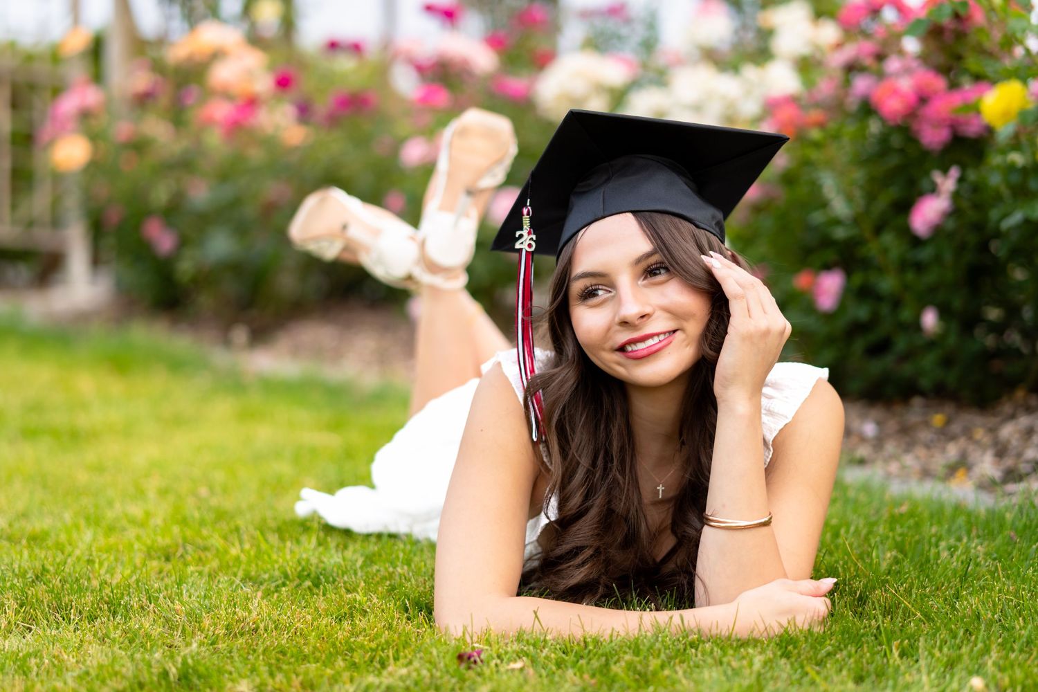 Smiling female graduate in cap lying on green grass with colorful roses blooming in the background.