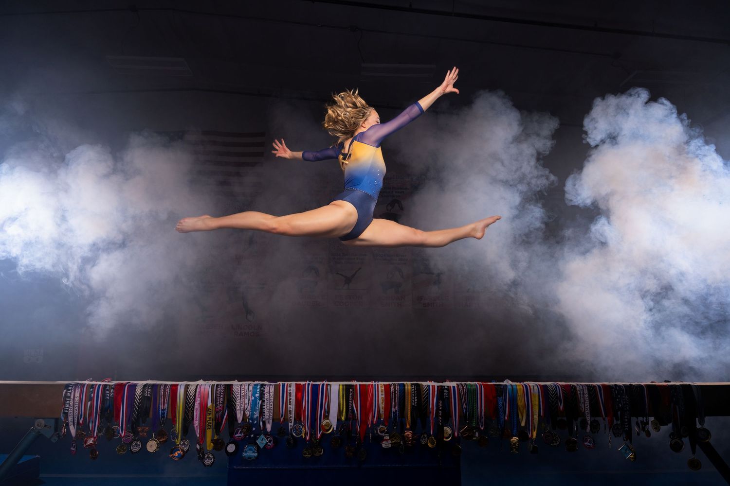 Young gymnast leaping mid-air in blue leotard surrounded by dramatic smoke, with colorful medals displayed below.