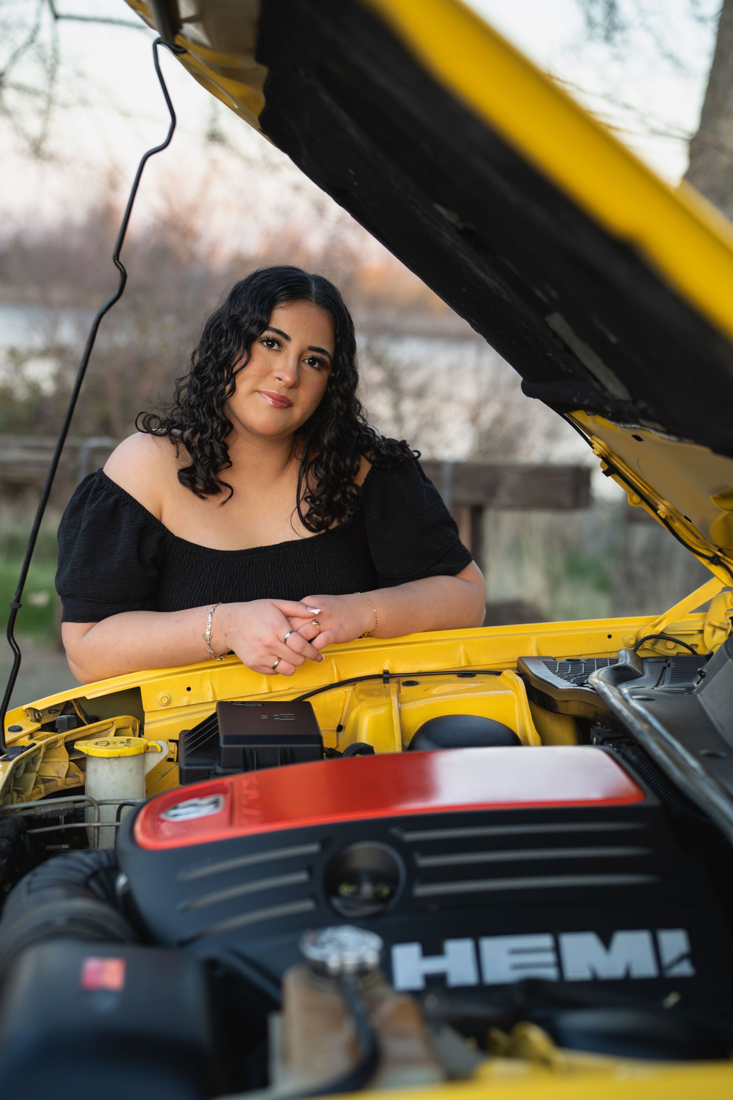 Young woman in black off-shoulder dress posing behind open hood of yellow Dodge HEMI muscle car outdoors.