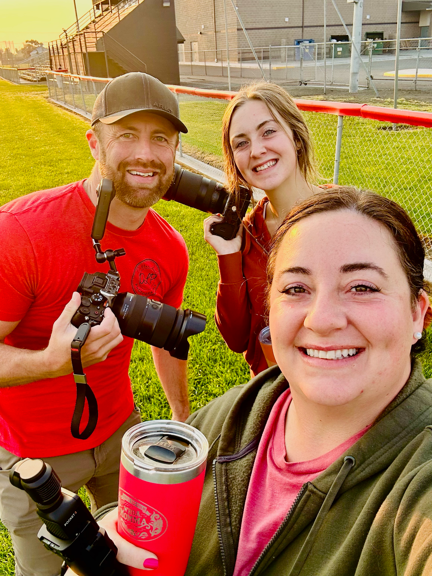 Three photographers with cameras smile together on a sunny field.