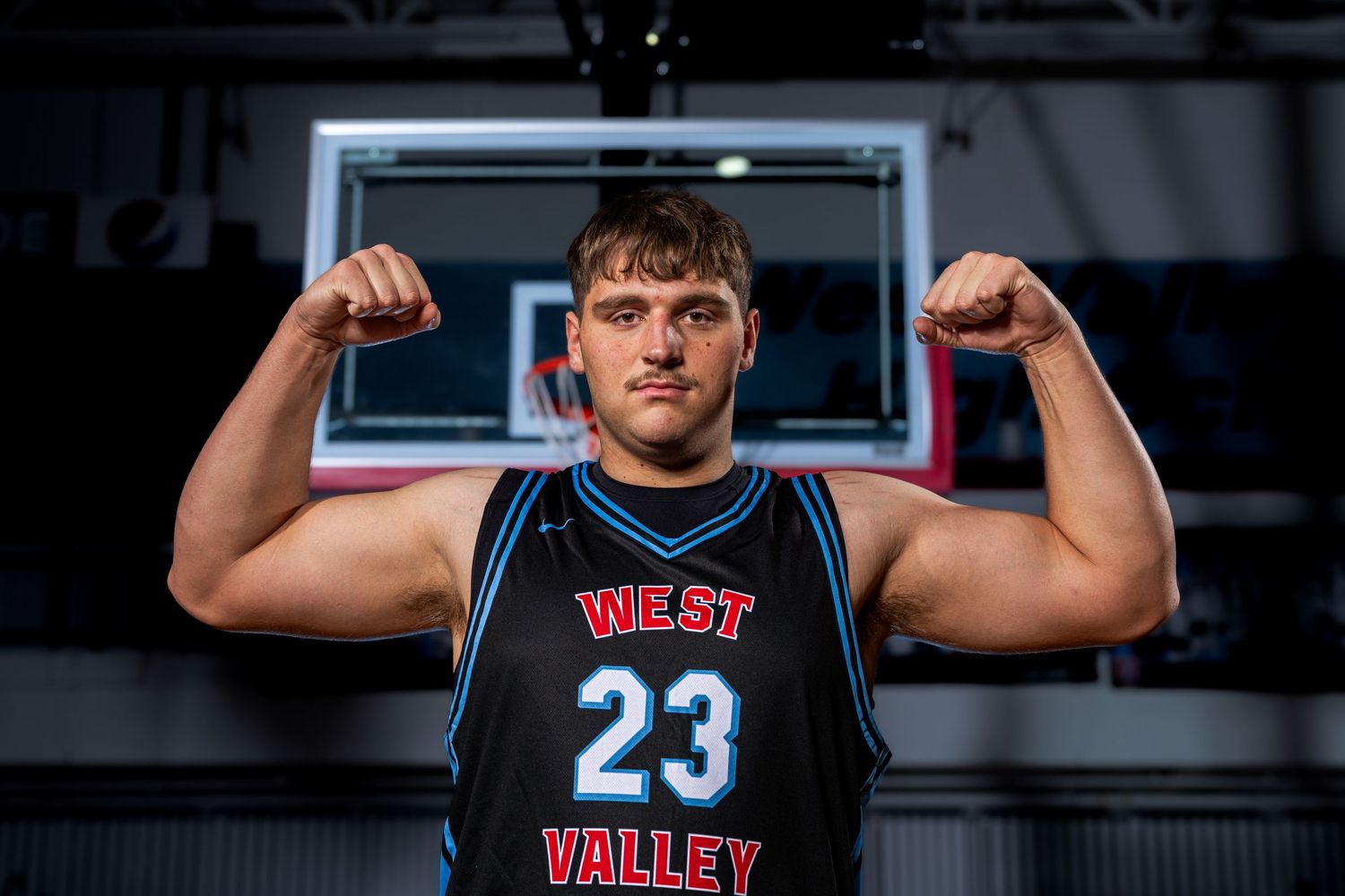 Basketball player in black West Valley jersey flexes muscles in front of backboard in indoor gym setting.
