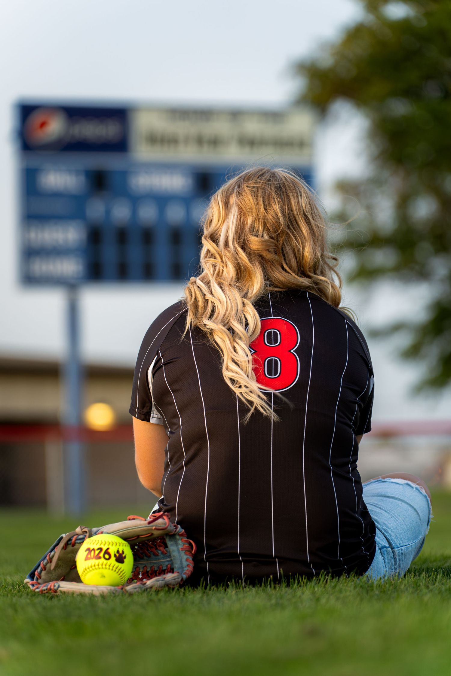 Softball player in jersey sits on grass field near scoreboard.