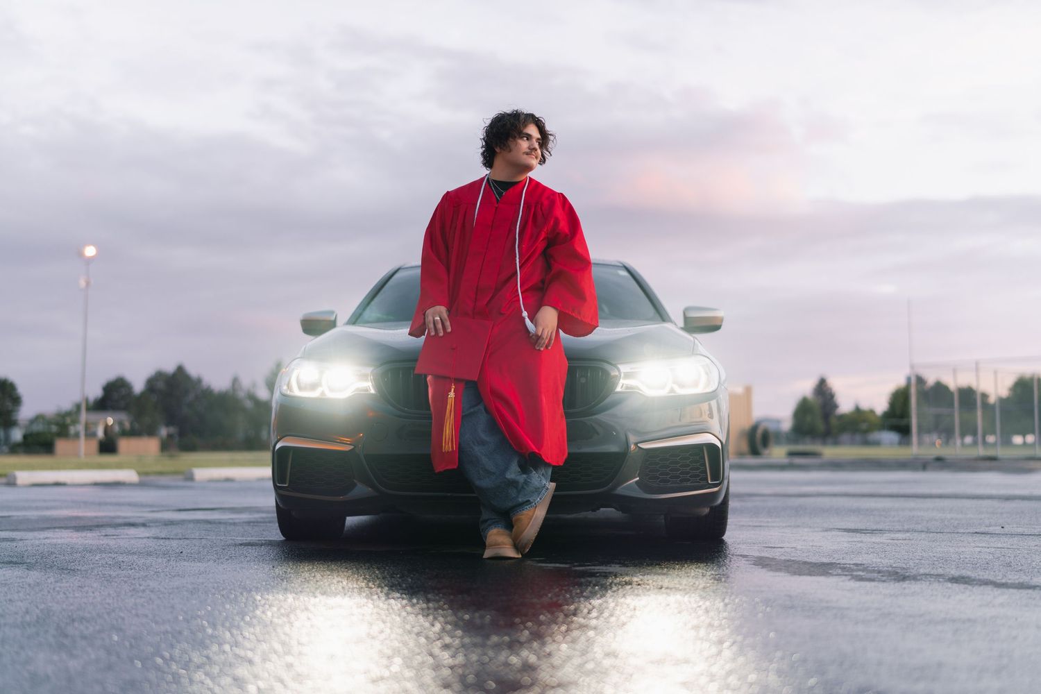 Person in red outfit leaning against a dark luxury car on wet pavement at dusk.