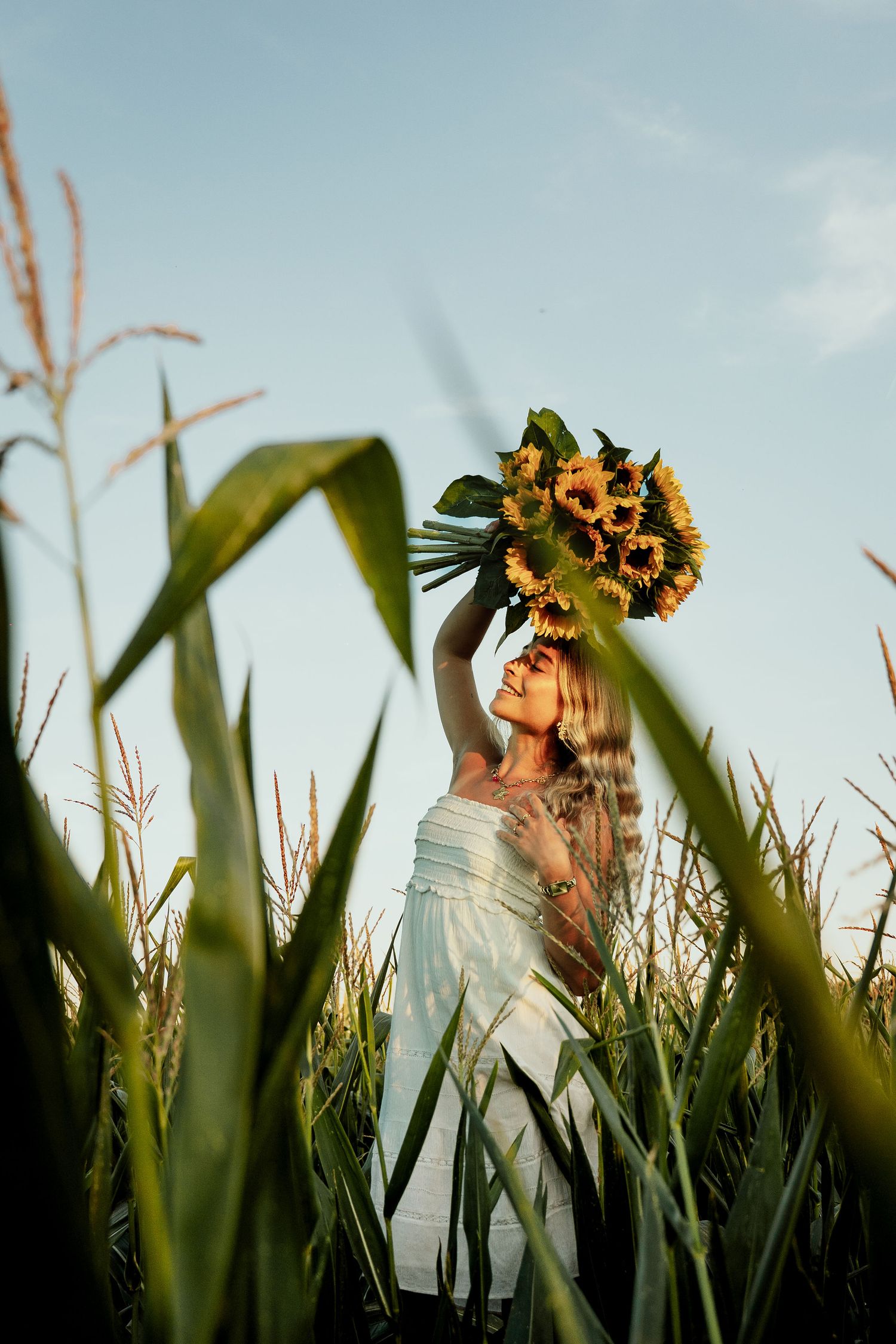 shooting photo d'une femme blonde portant une robe blanche dans un champ de mais avec un bouquet de tournesols