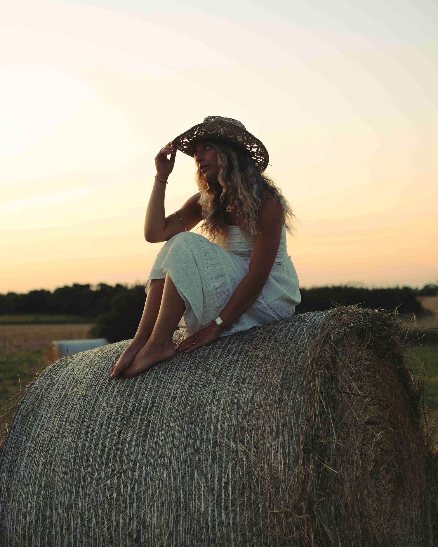 modele photo habillée en blanc avec un chapeau en paille assise sur une botte de foin