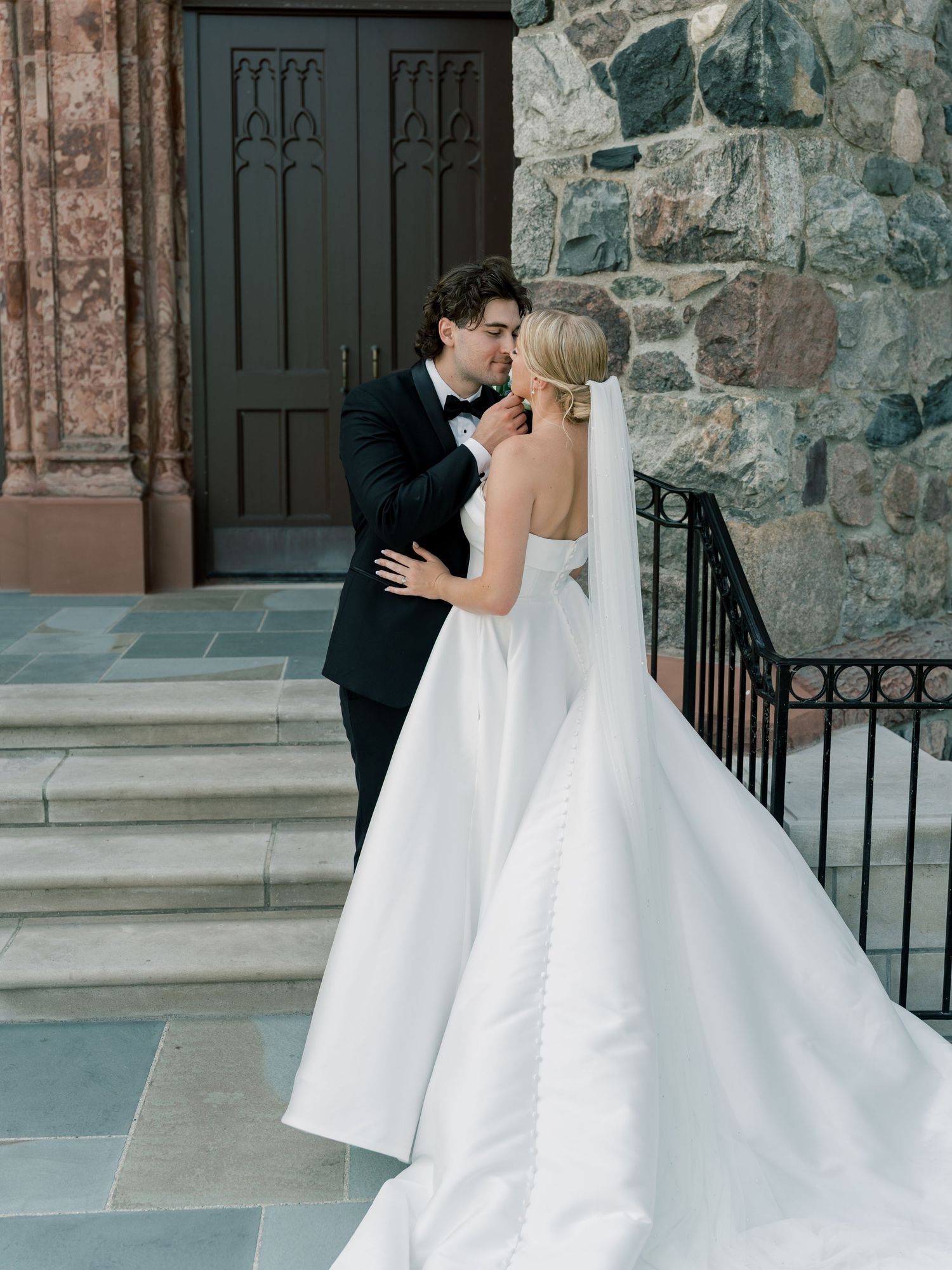 Bride in white ballgown and groom in tuxedo share a kiss on stone church steps.