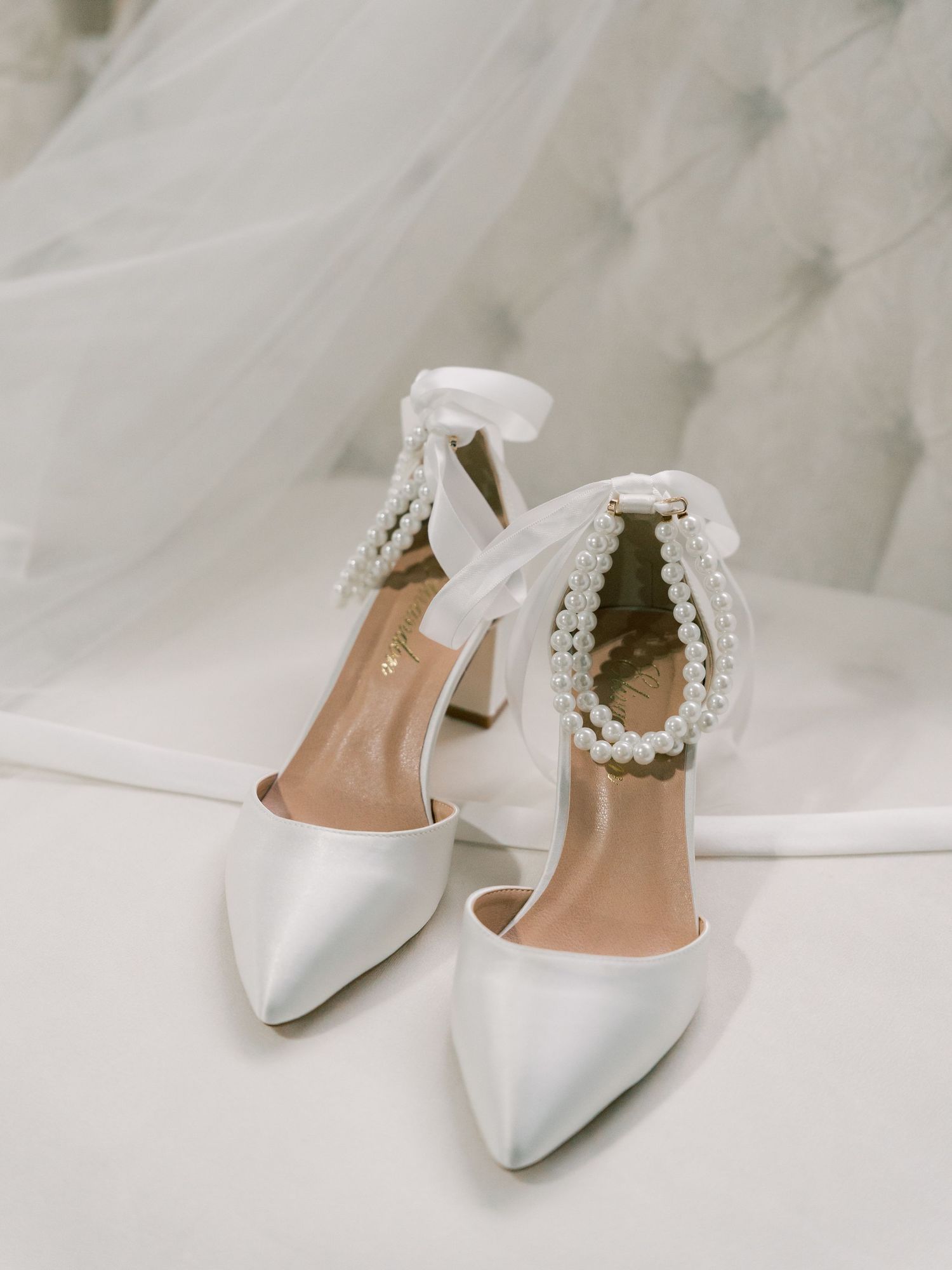 Elegant white satin bridal heels with pearl embellished ankle straps displayed on marble background.