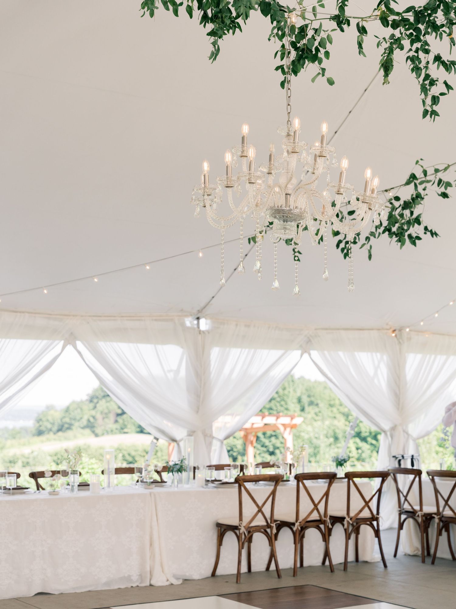 Elegant white wedding tent interior with chandeliers, string lights and wooden cross-back chairs set up for reception.