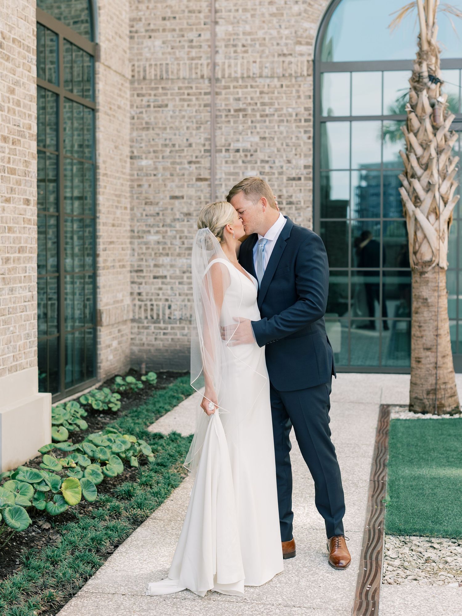 A romantic wedding couple shares an intimate moment outside a brick building with palm trees and arched windows.