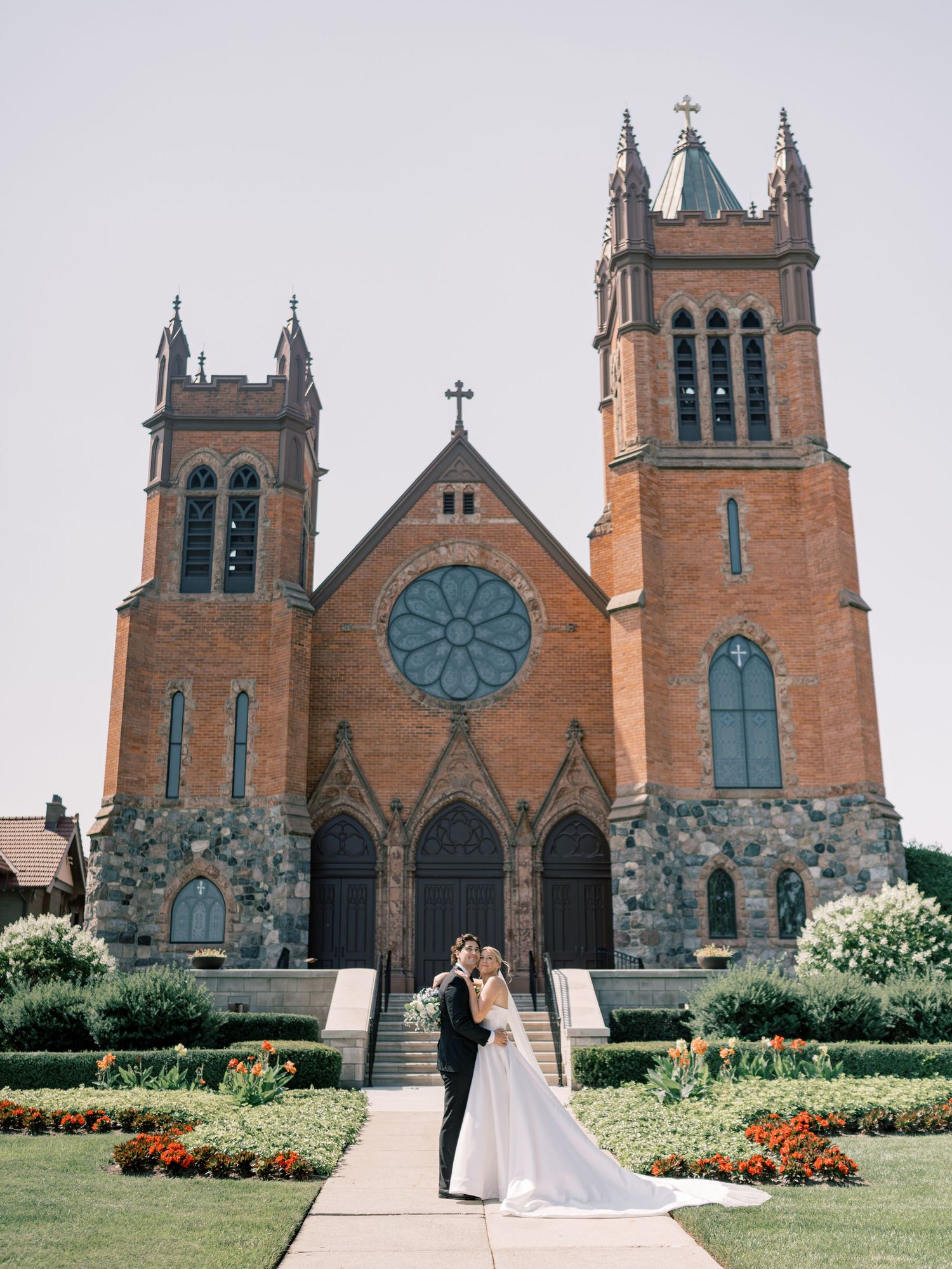 Bride and groom embrace before a grand red brick Gothic church with rose window and twin towers.