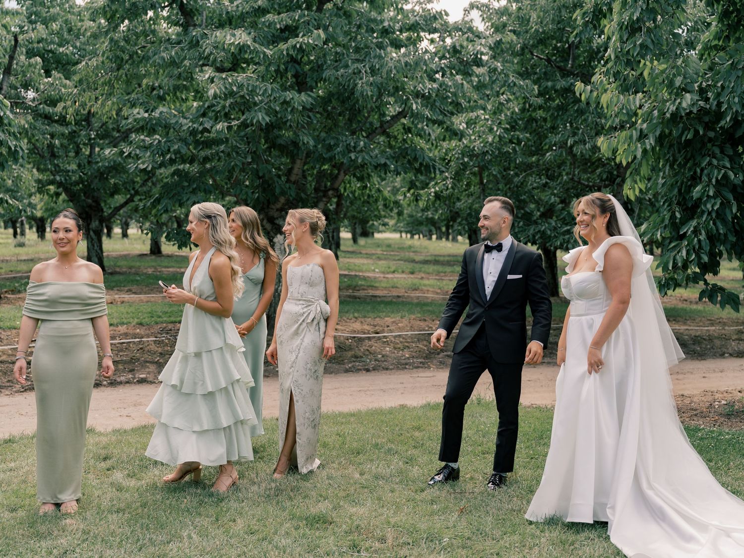 Wedding party members walk through an orchard during a summer celebration in elegant formal attire.