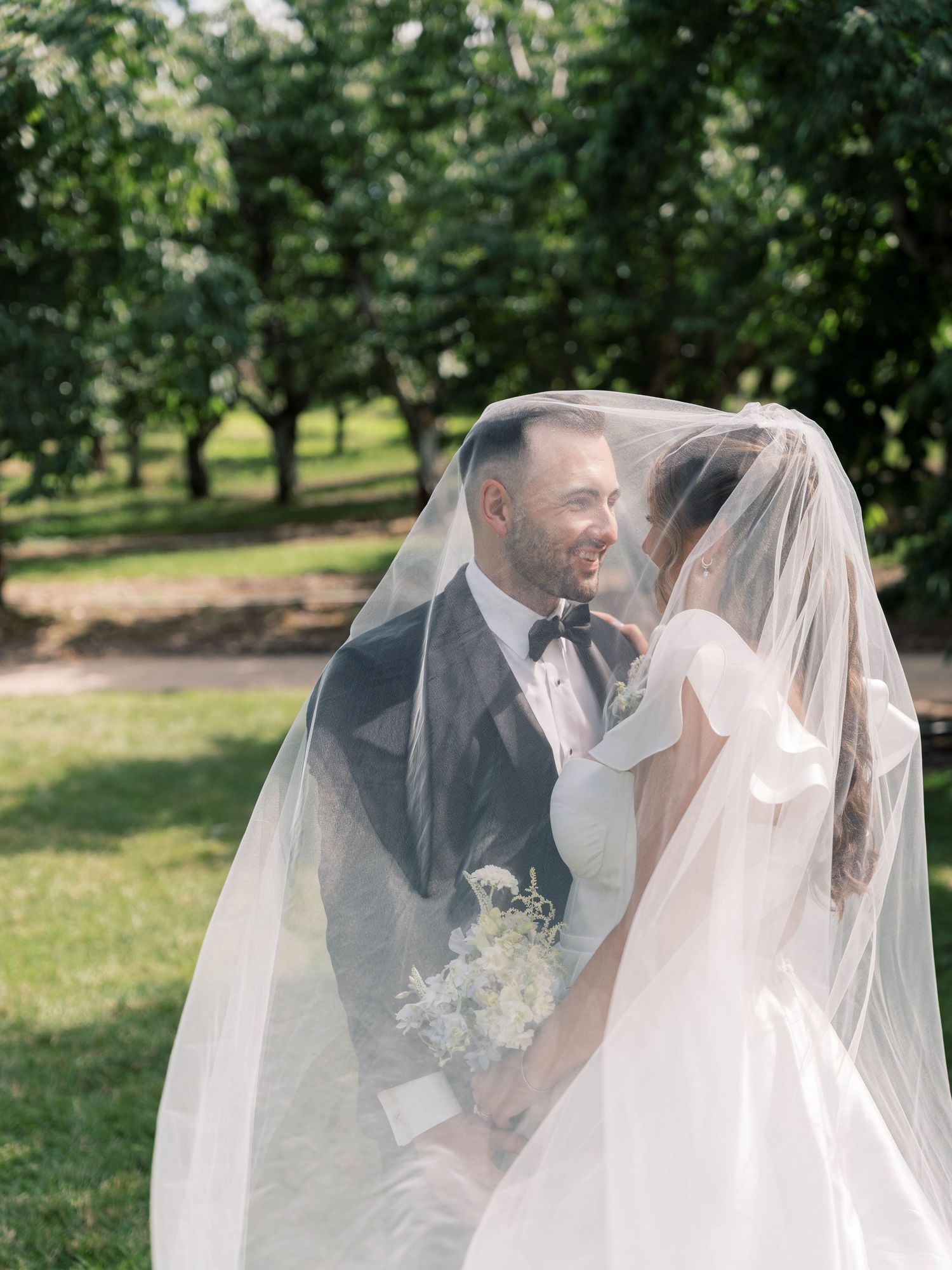 Newlyweds share an intimate moment under a flowing veil in a sunlit garden setting.