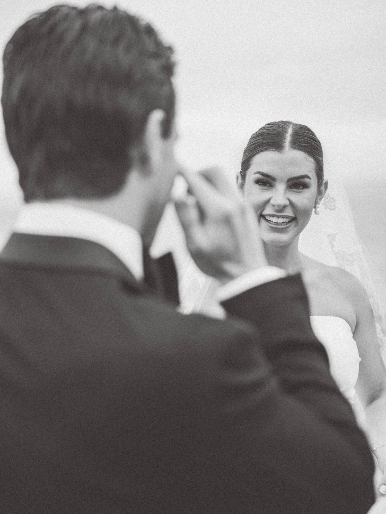 Black and white wedding photo of a groom wiping a tear from his smiling bride's face during the ceremony.