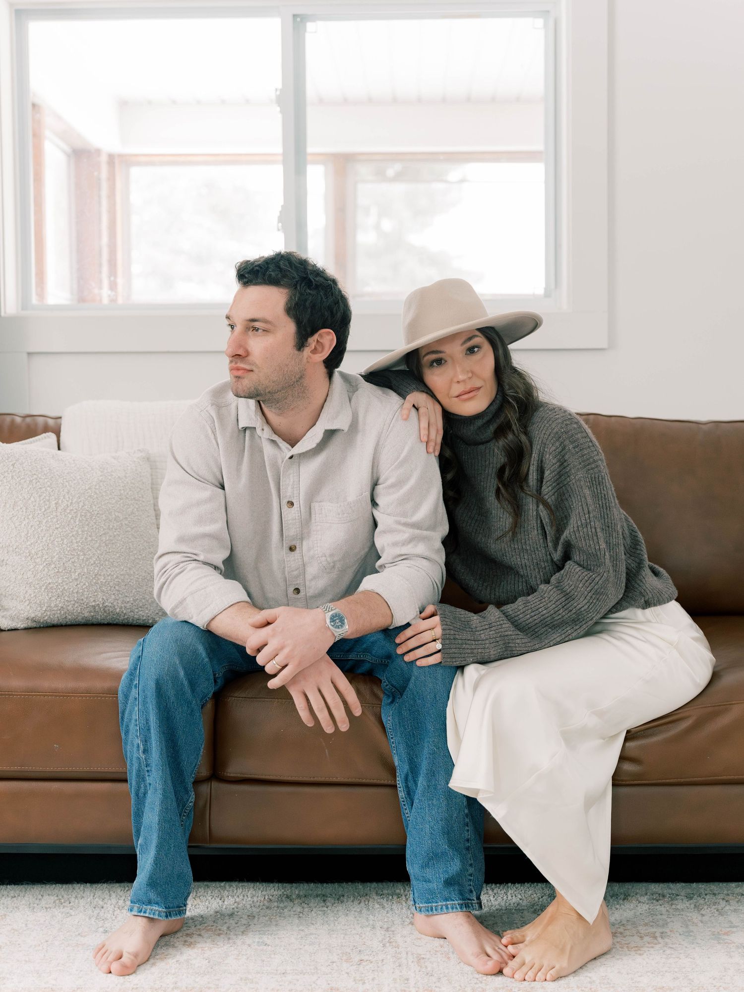 A couple lounges casually on a brown leather couch in a bright, minimalist living room setting.