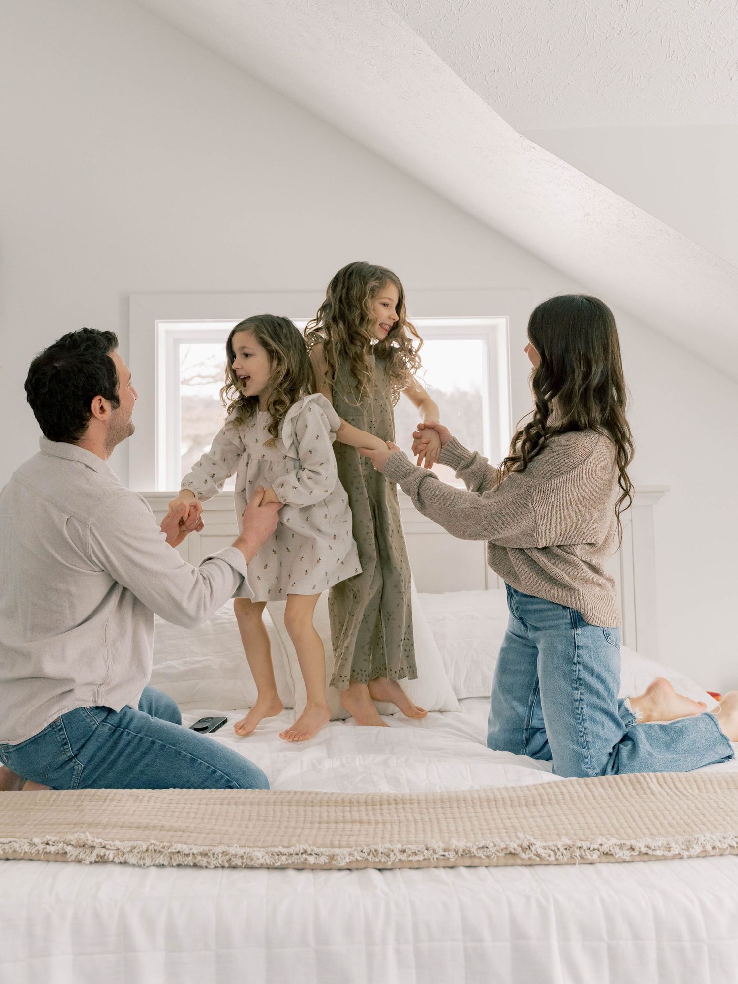 A family enjoying playful moments together while jumping on a bed in a bright white room with natural lighting.
