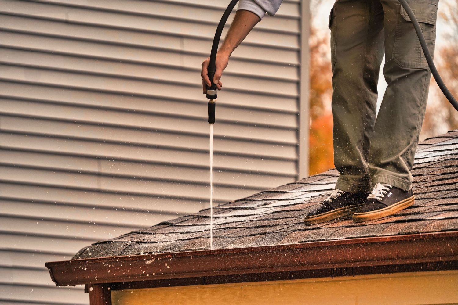 Using a pressure washer to clean dirt and debris off of a roof with vinyl siding visible in the background.