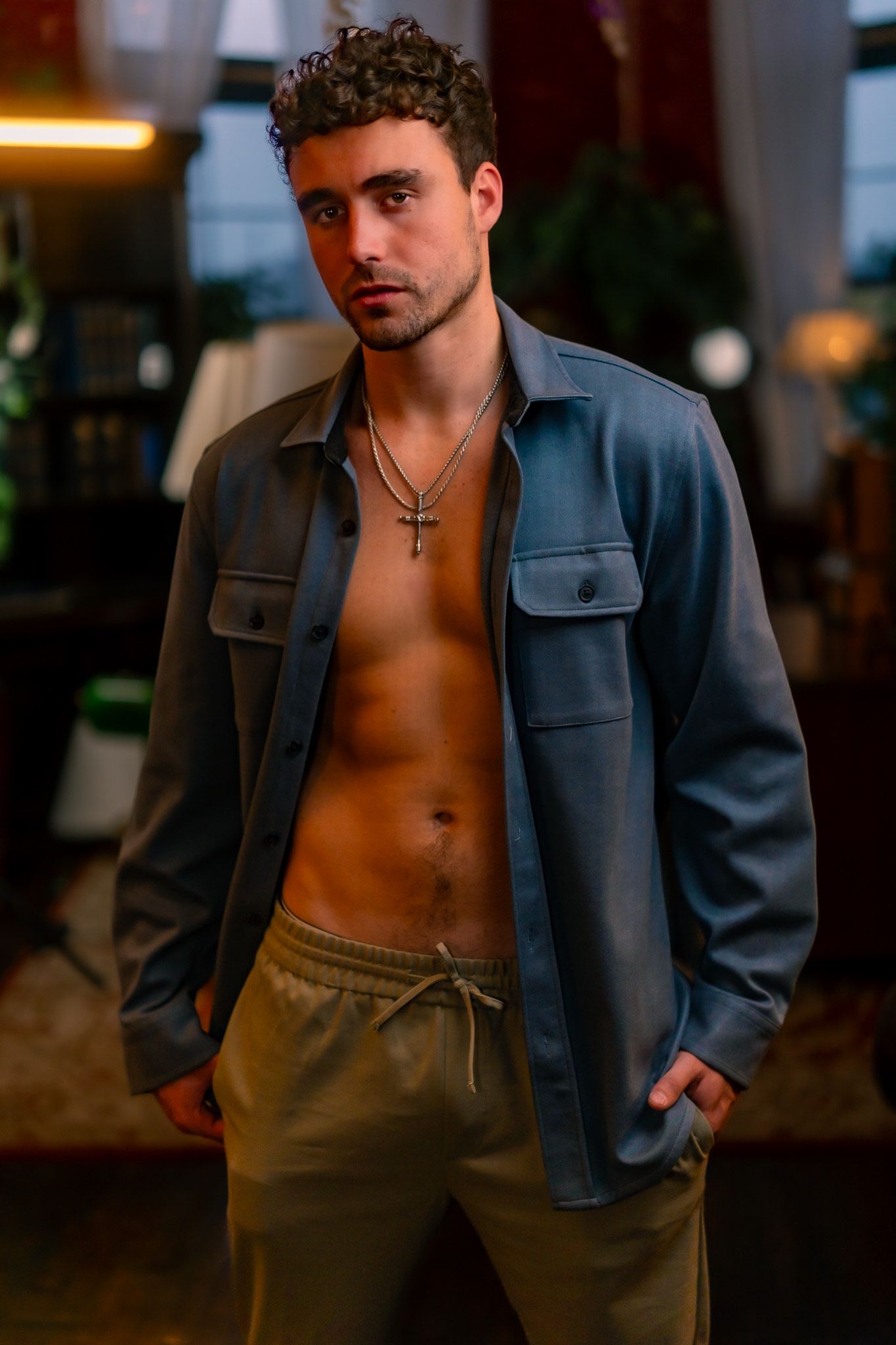 A person wearing an open denim jacket and tan sweatpants poses in moody indoor lighting with a necklace.