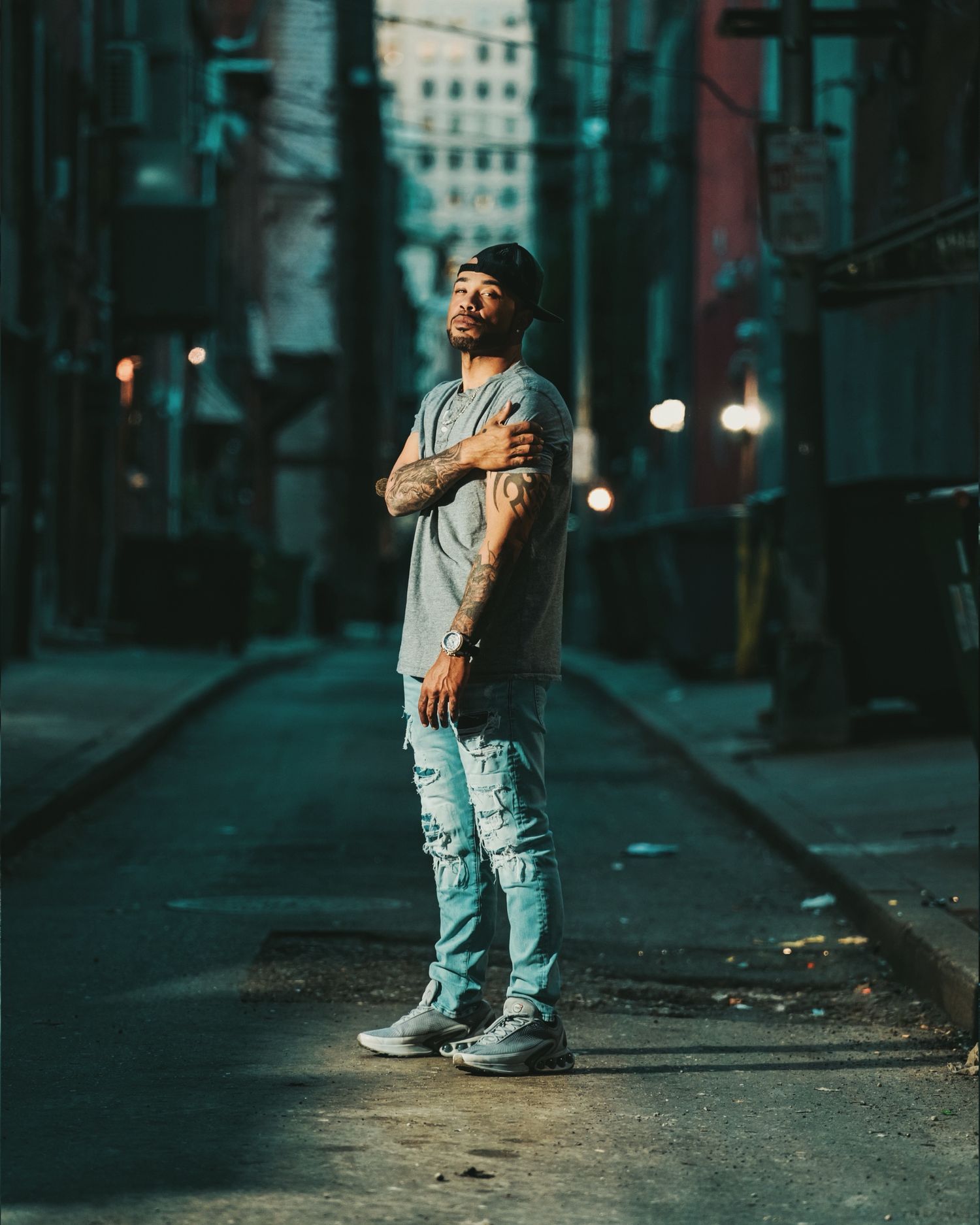 Person in ripped jeans and gray shirt poses confidently on moody urban street at night with bokeh lighting effects.