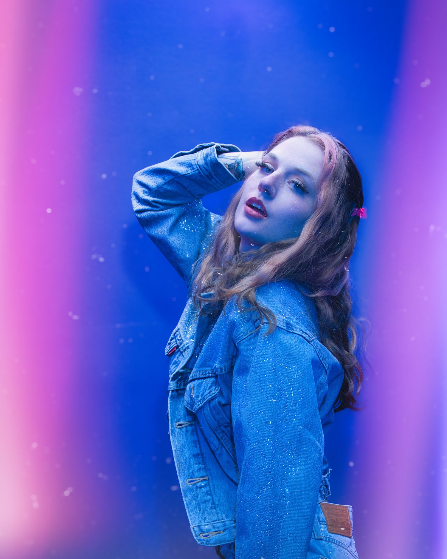 Artistic portrait with denim jacket against vibrant blue and pink lighting with dreamy sparkle effects in background.