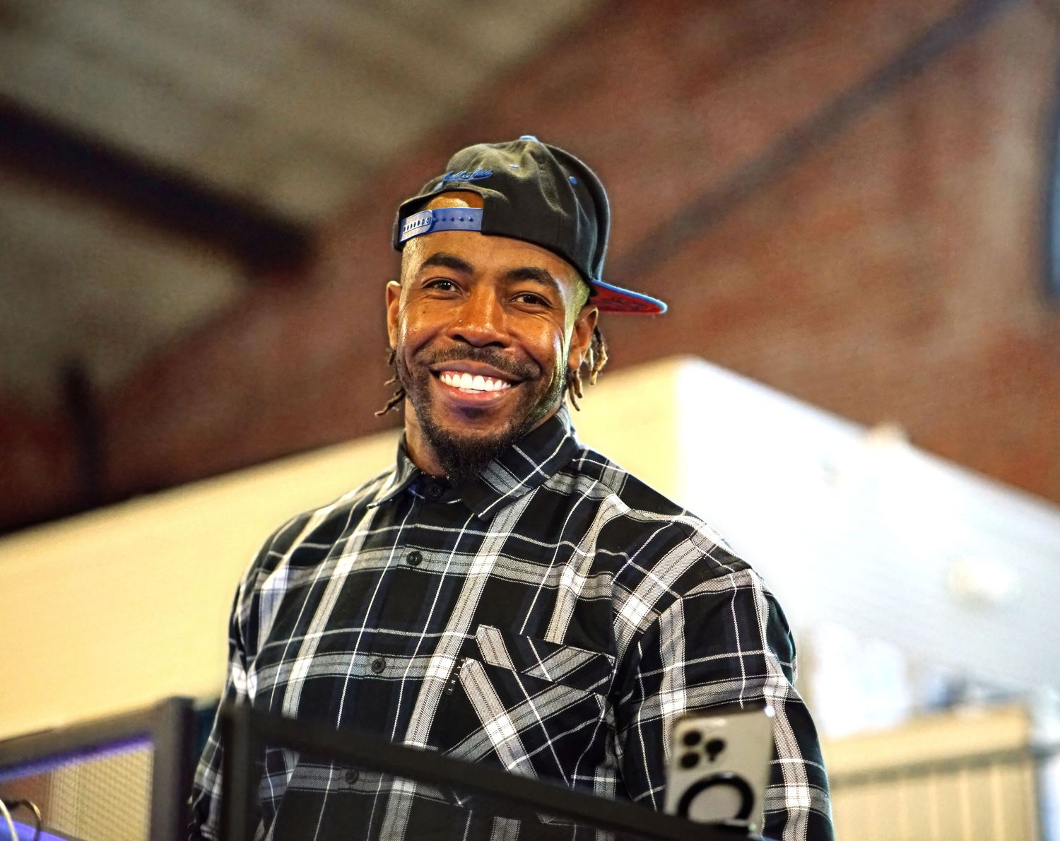 A person wearing a backwards baseball cap and plaid flannel shirt smiles warmly against a blurred brick wall background.