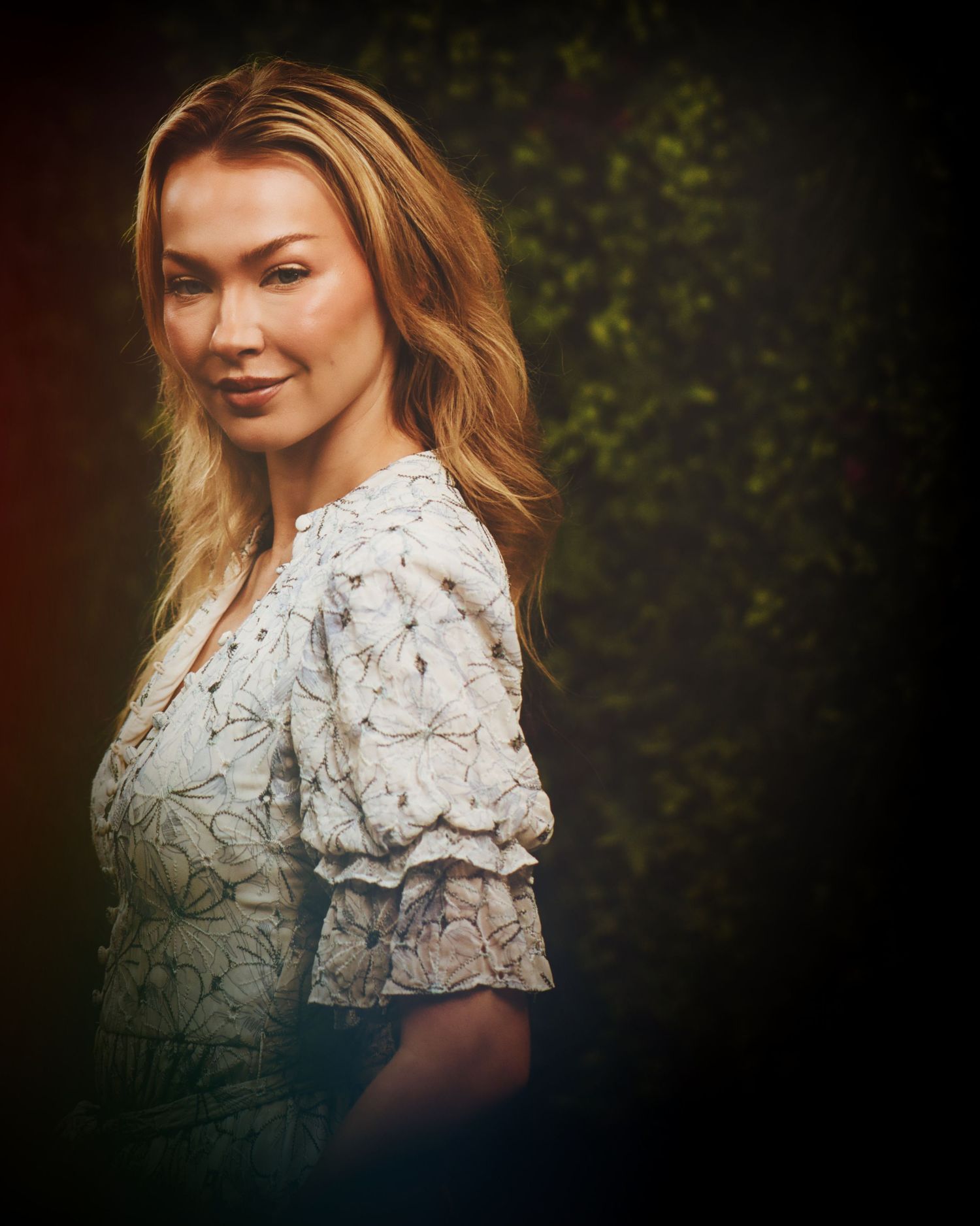 Portrait of someone wearing a white lace blouse with blonde highlighted hair against a dark nature backdrop.