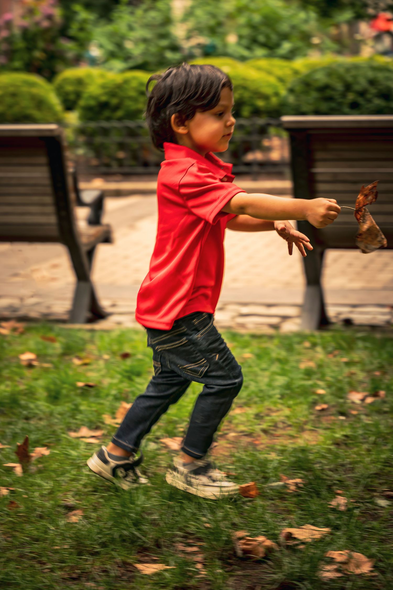 A child in a red shirt and jeans jumps playfully on grass covered with autumn leaves in a park setting.
