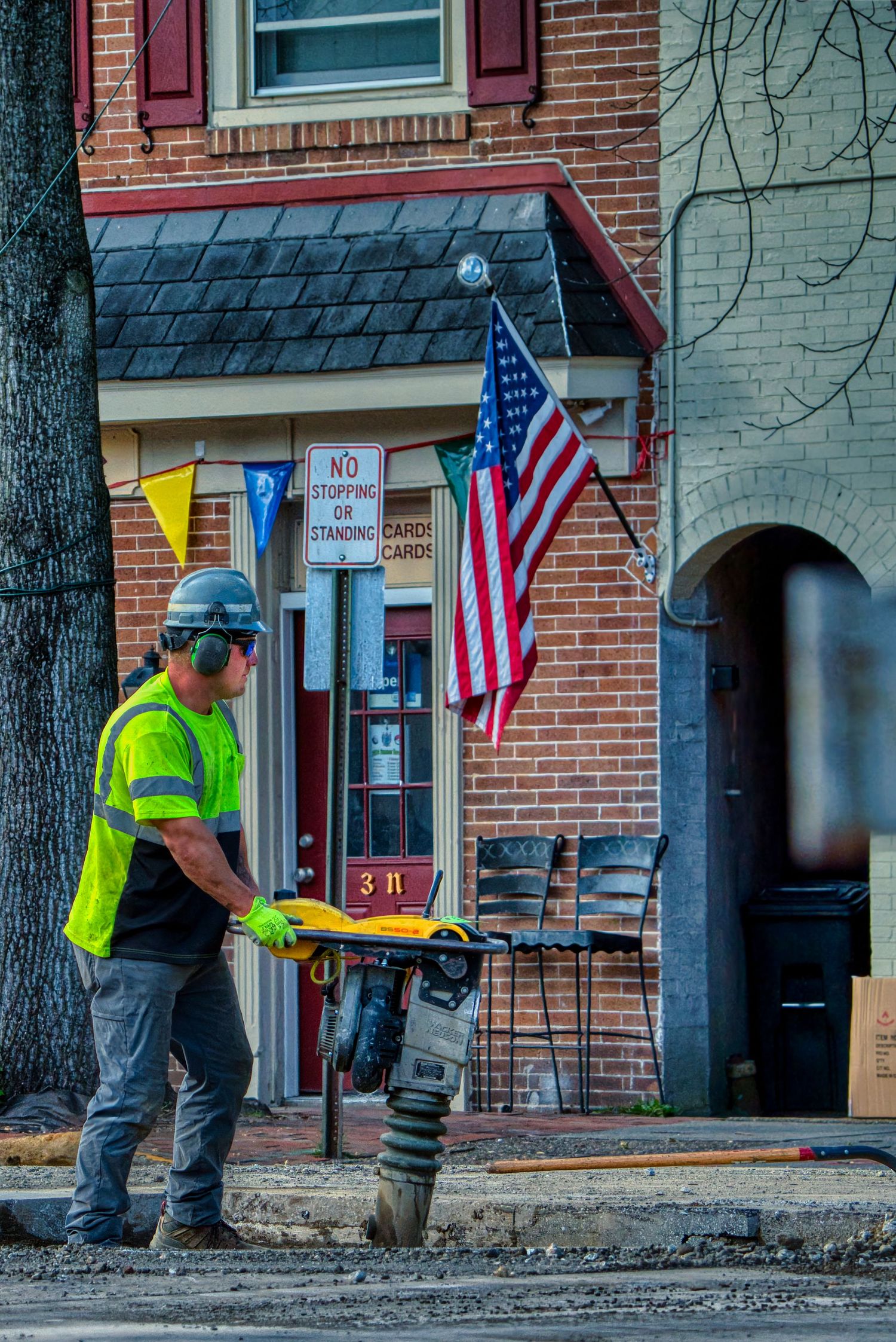 Construction worker in safety vest operates a soil compactor on a street with an American flag visible in background.