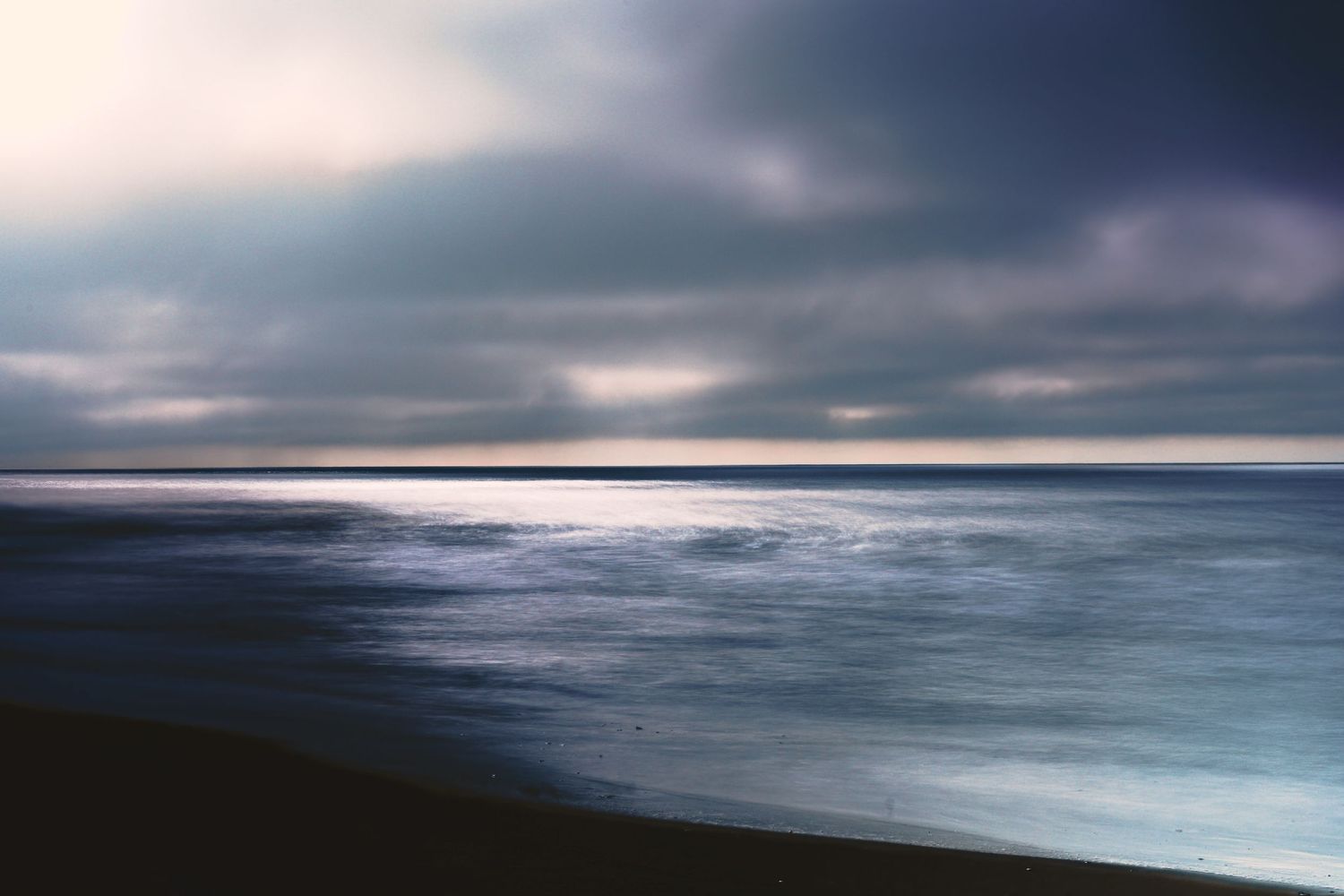 Dramatic ocean landscape at dusk with stormy clouds reflected in dark rolling waves against a mountainous coastline.