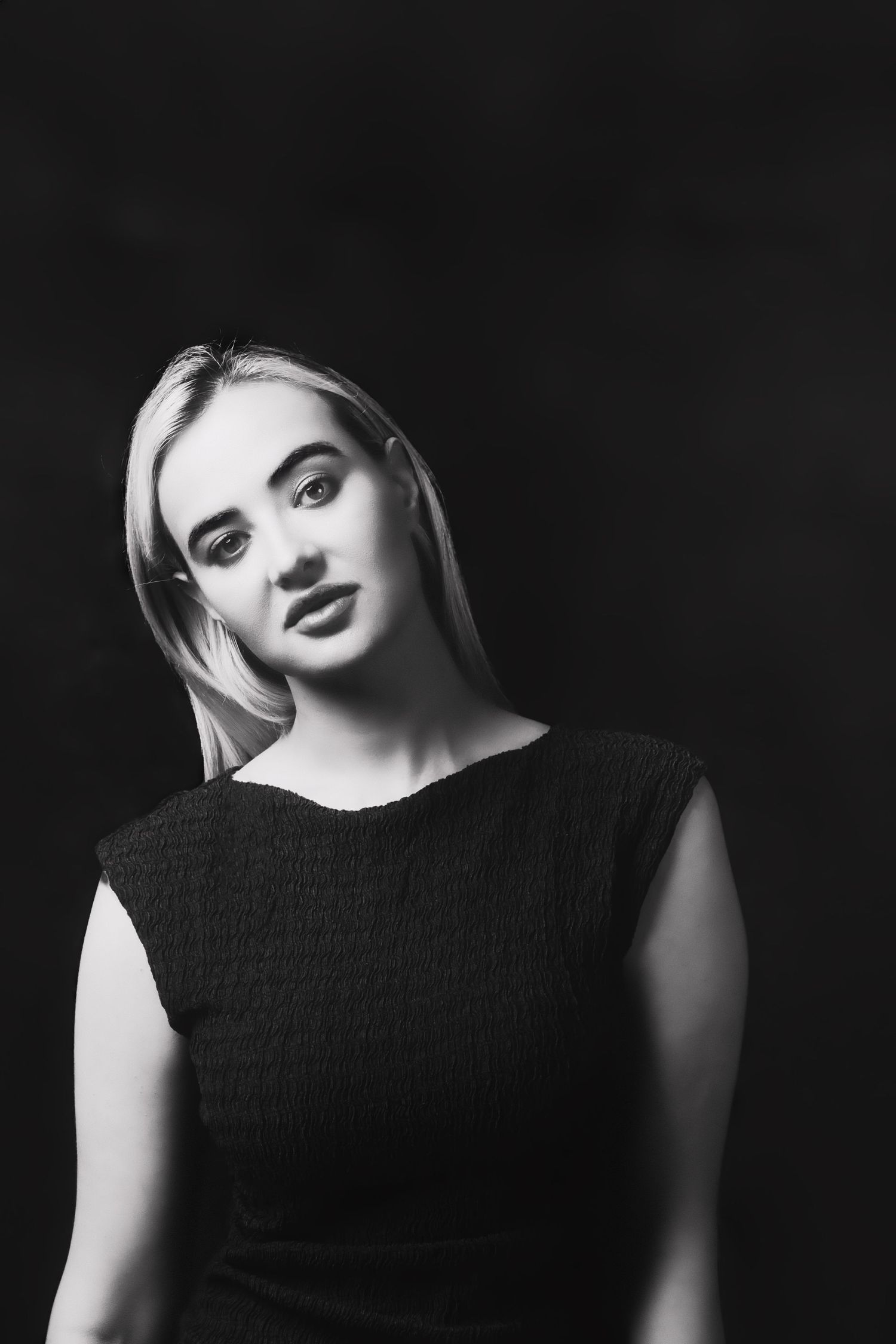 Black and white portrait photograph featuring a blonde woman in a black sleeveless top against a dark background.
