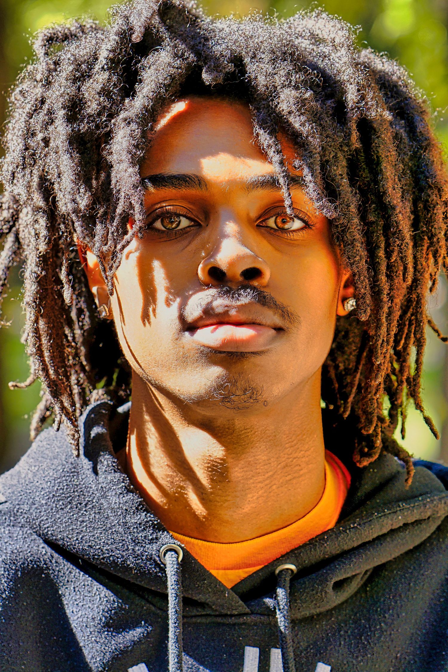 Person wearing a dark hoodie with dreadlocks sits against a blurred green outdoor background, taken in close-up.