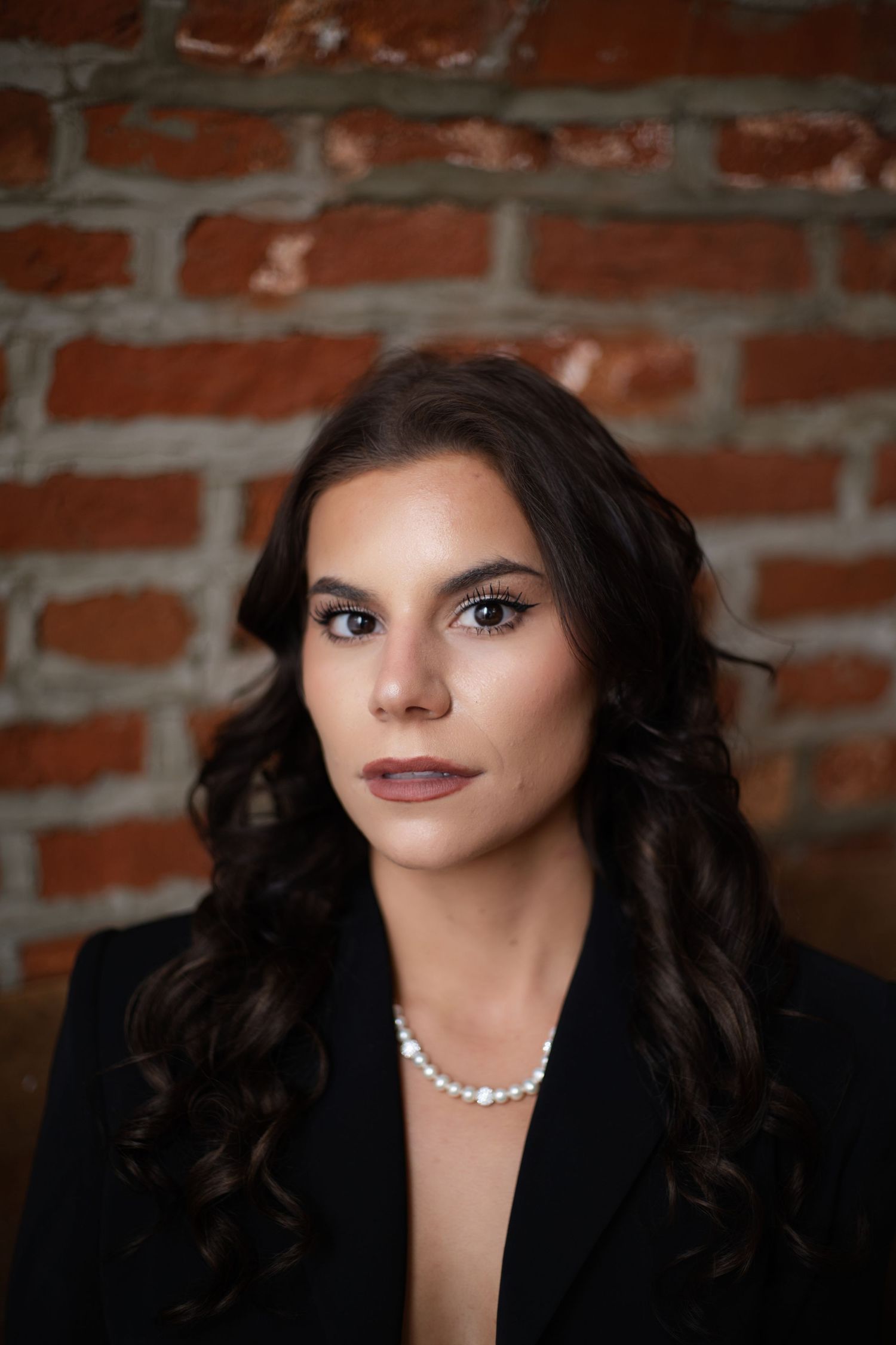 Professional portrait against brick wall featuring dark curly hair, pearl necklace and dramatic makeup.