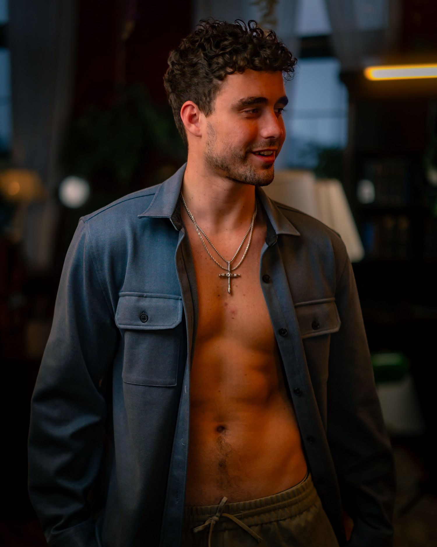 A person wearing an open denim jacket showing bare chest and torso stands indoors in warm lighting.