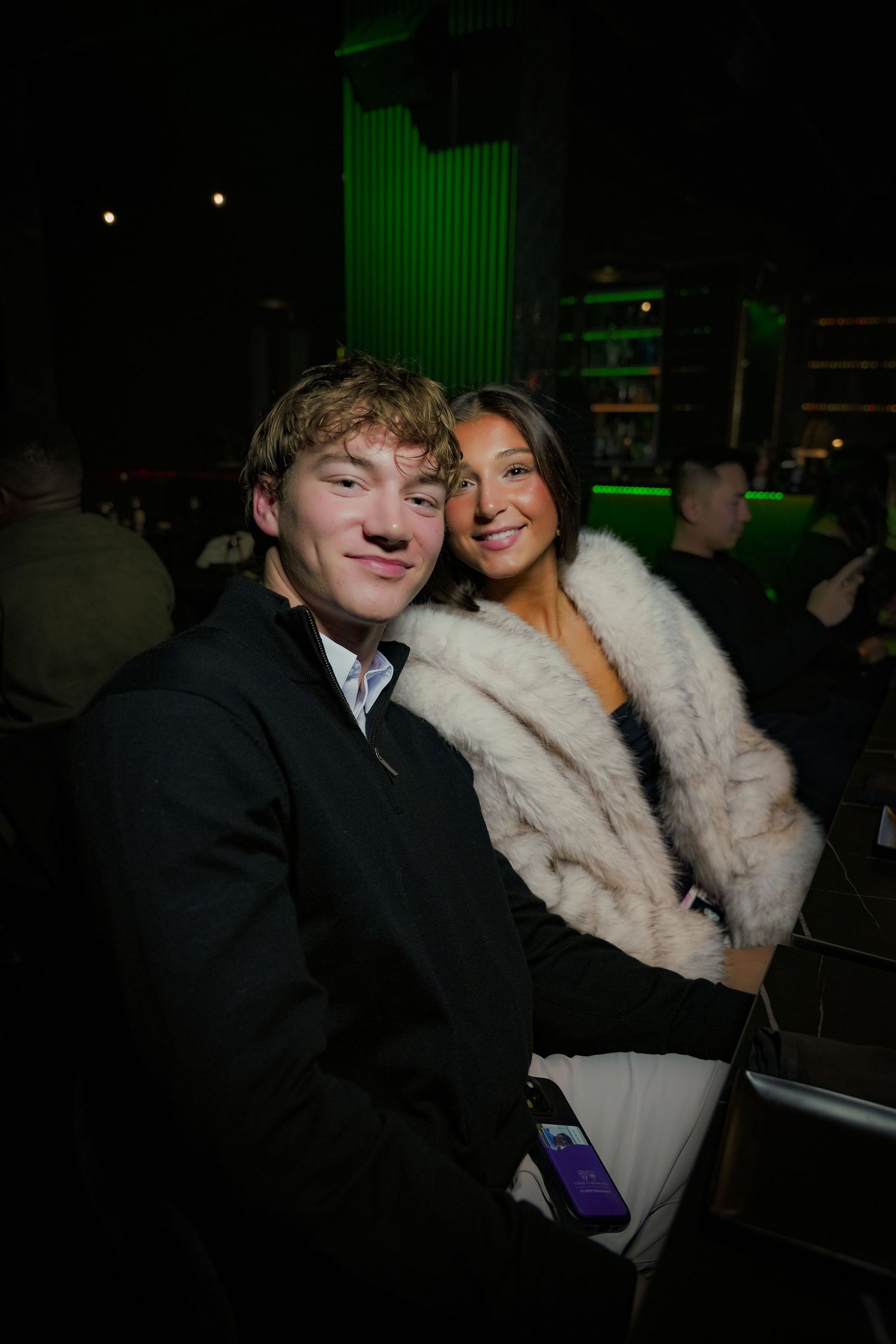 Two people pose together in a dimly lit venue with green lighting visible in the background.
