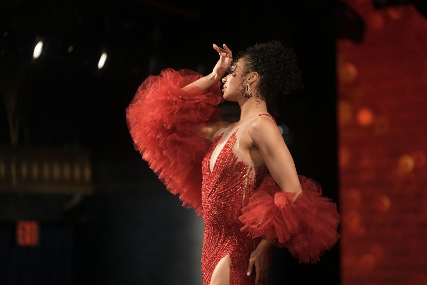 A performer in a red sequined costume with dramatic ruffles strikes a pose on stage during a theatrical show.