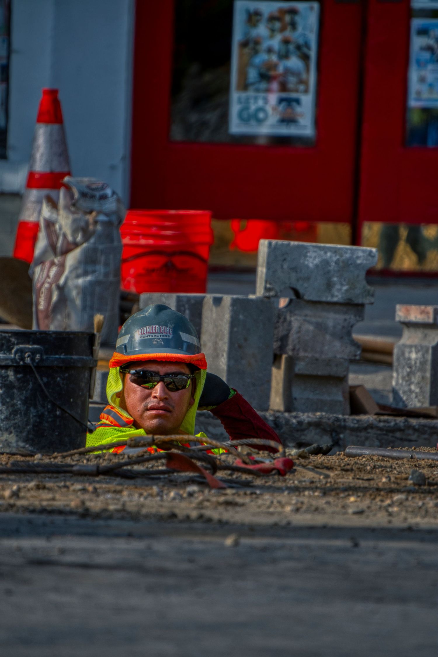 Construction worker in high visibility gear and dark sunglasses peering up from below ground level at a worksite.