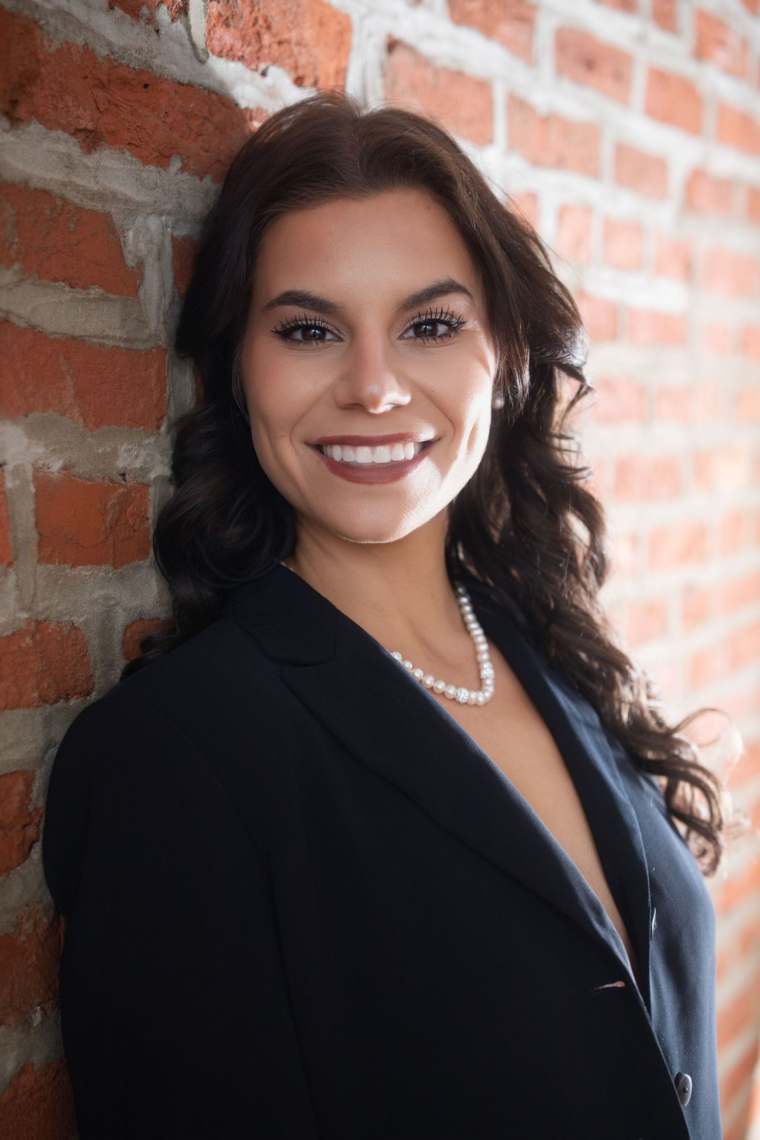 Professional headshot against exposed brick wall featuring dark blazer, pearl necklace and warm smile.