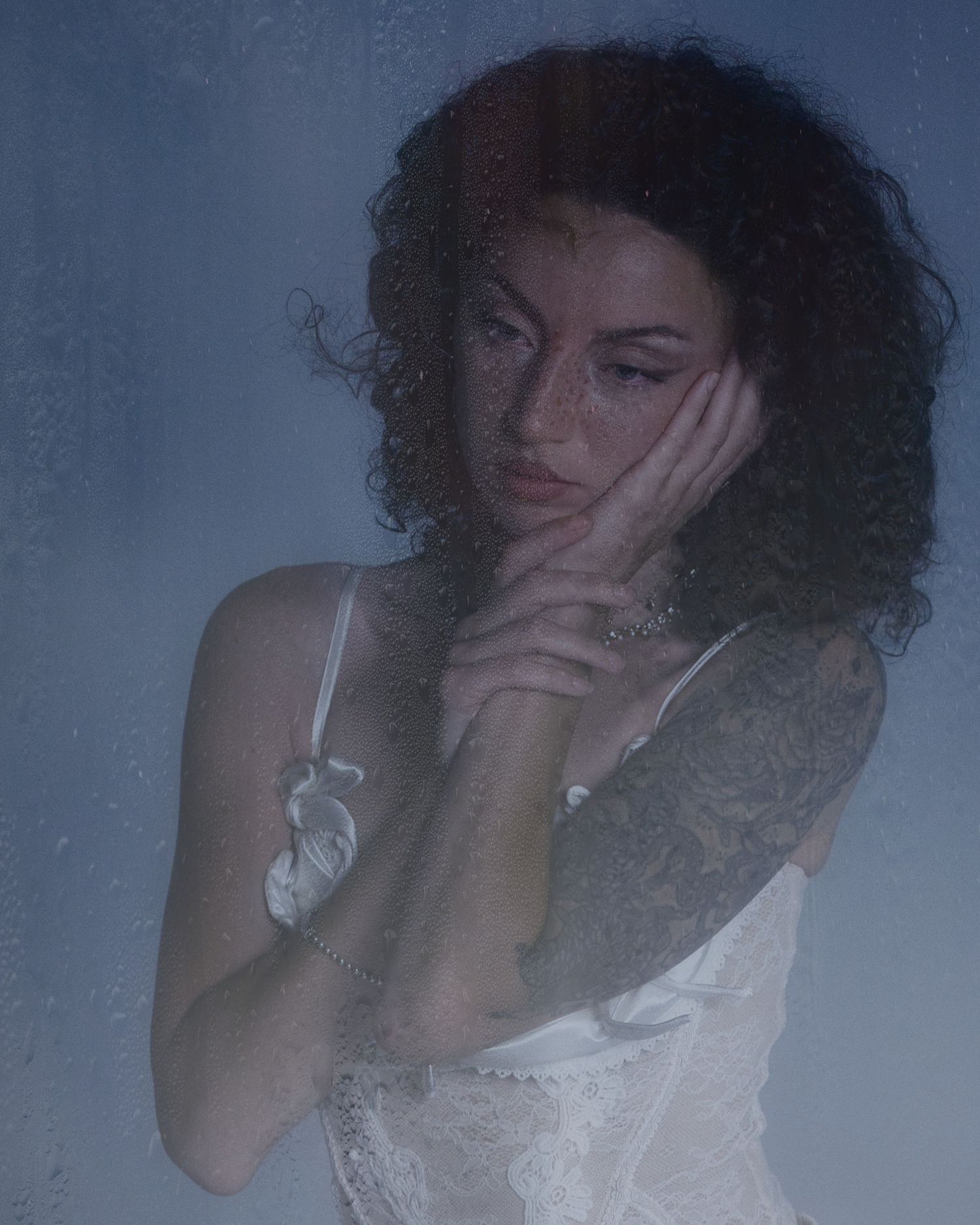 A person with curly dark hair wearing a white lace top poses in a dreamy, foggy studio setting.