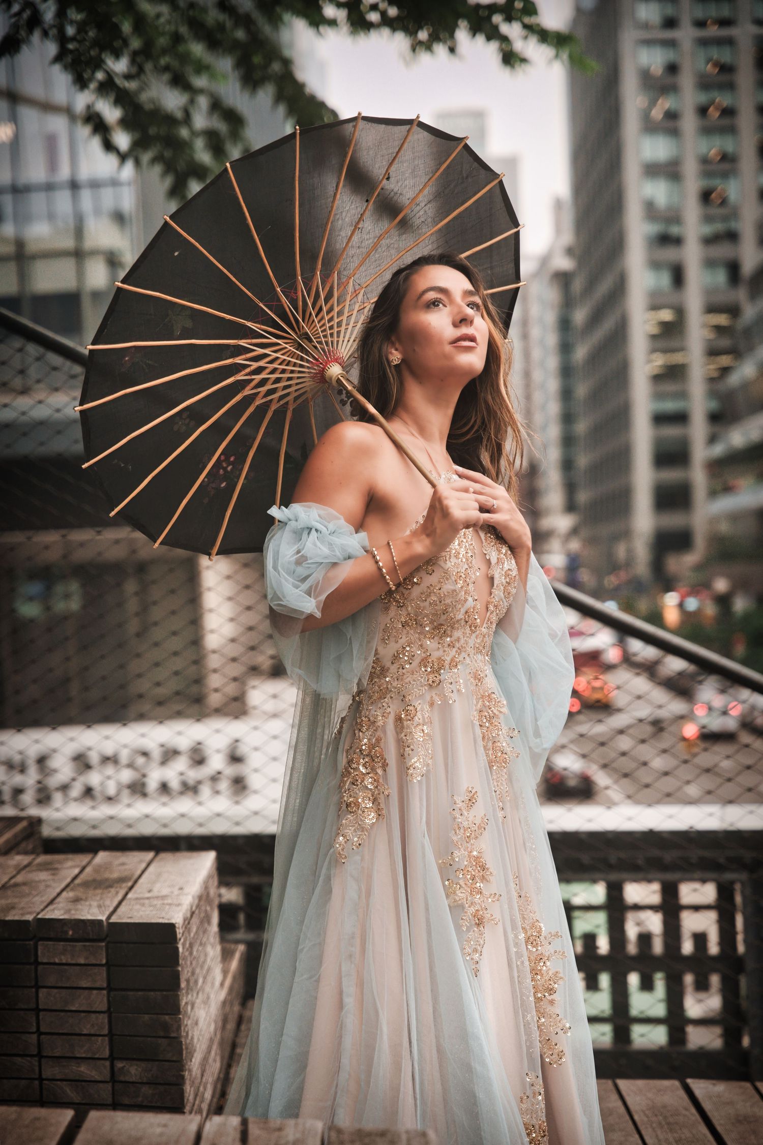 Elegant fashion portrait featuring an ornate parasol and flowing embellished gown against an urban cityscape backdrop.
