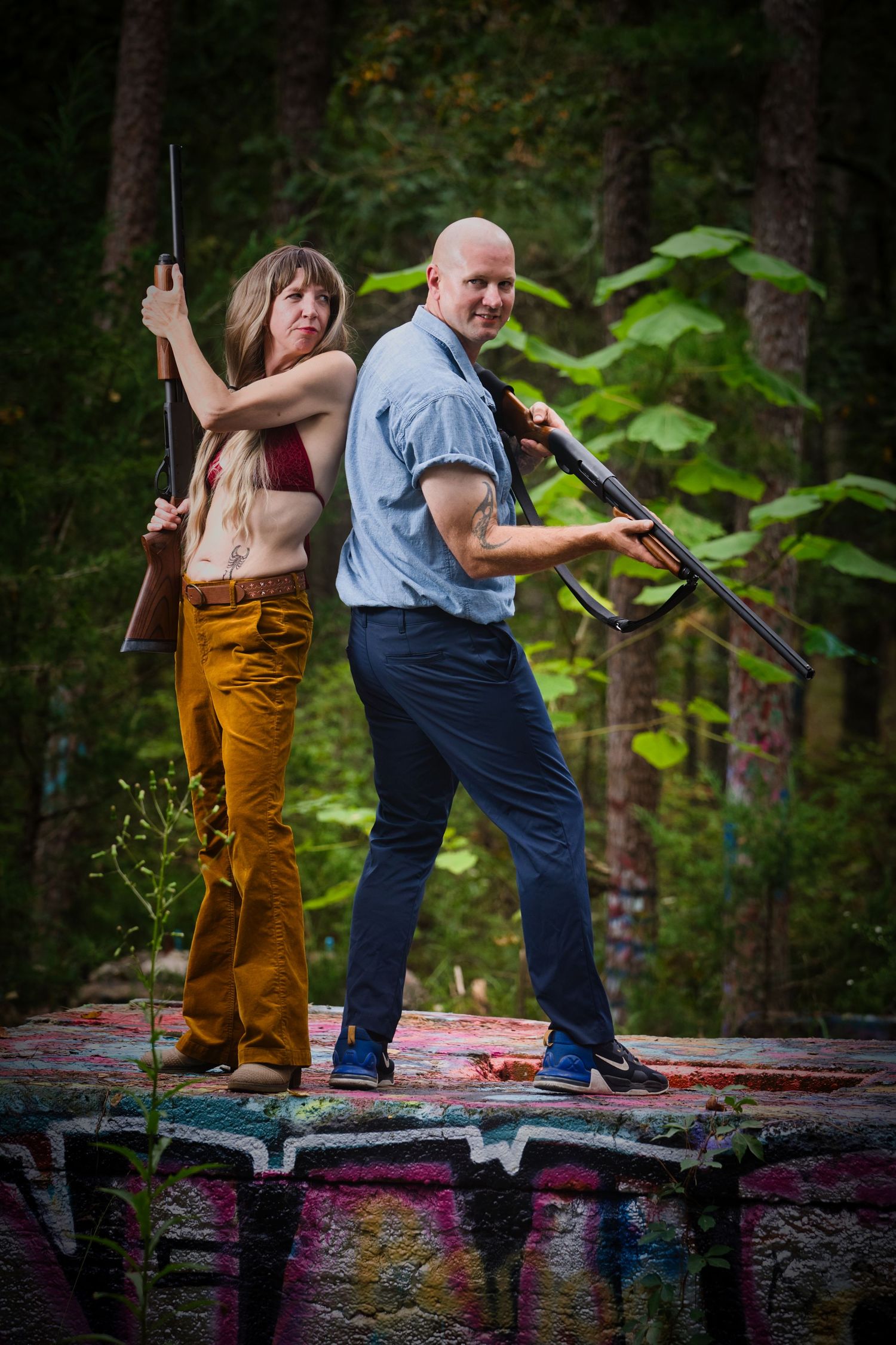 Two people stand back-to-back holding rifles on a colorful graffiti-covered concrete surface in a forest setting.
