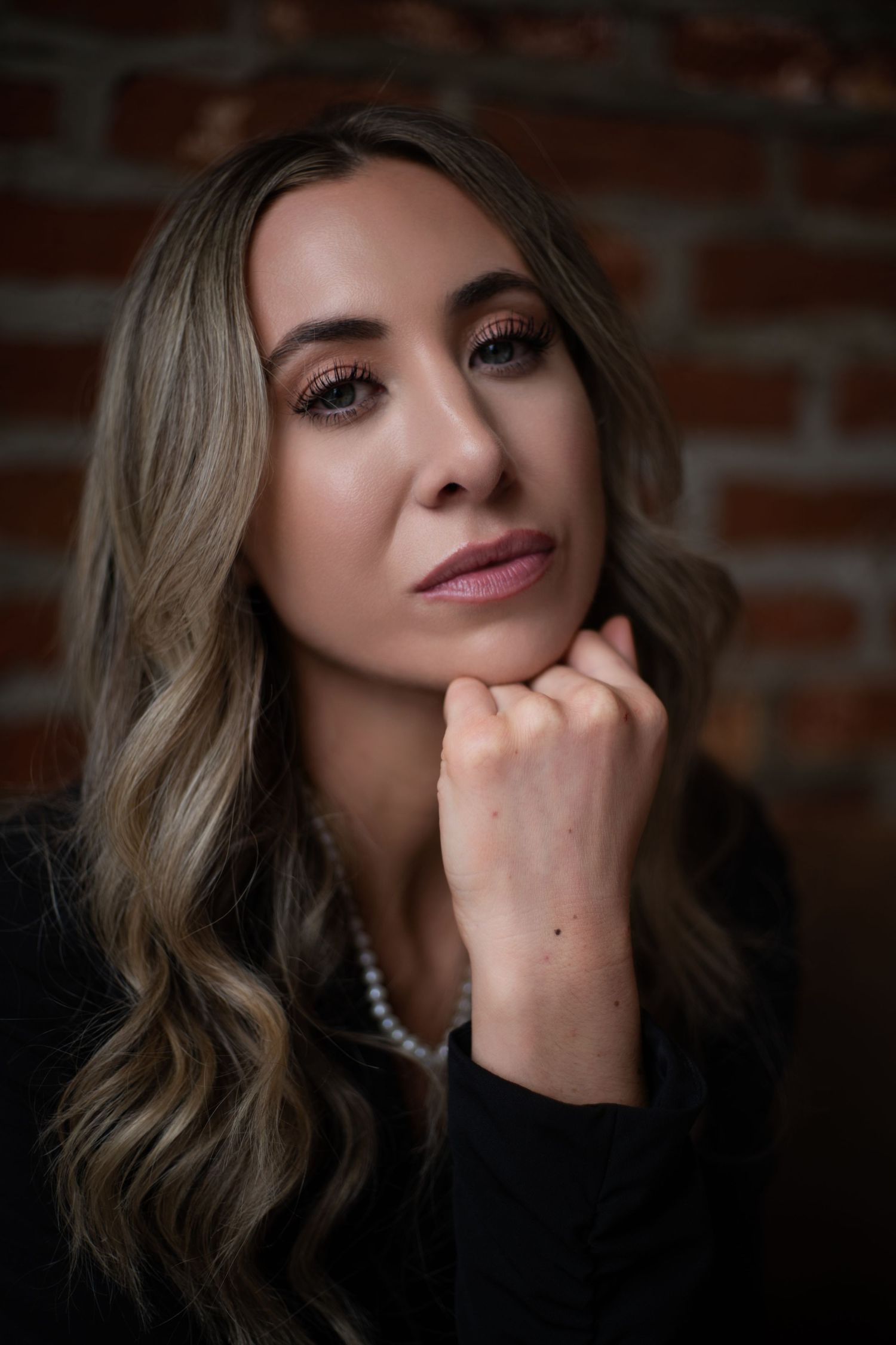 A series of moody portrait photographs showing soft lighting, wavy hair, and pearl necklace details against a brick wall backdrop.
