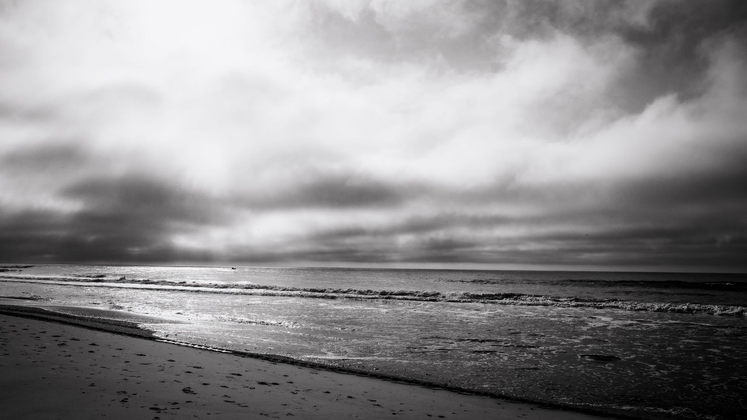 A dramatic black and white panorama of ocean waves breaking on a beach under stormy clouds stretches across the horizon.
