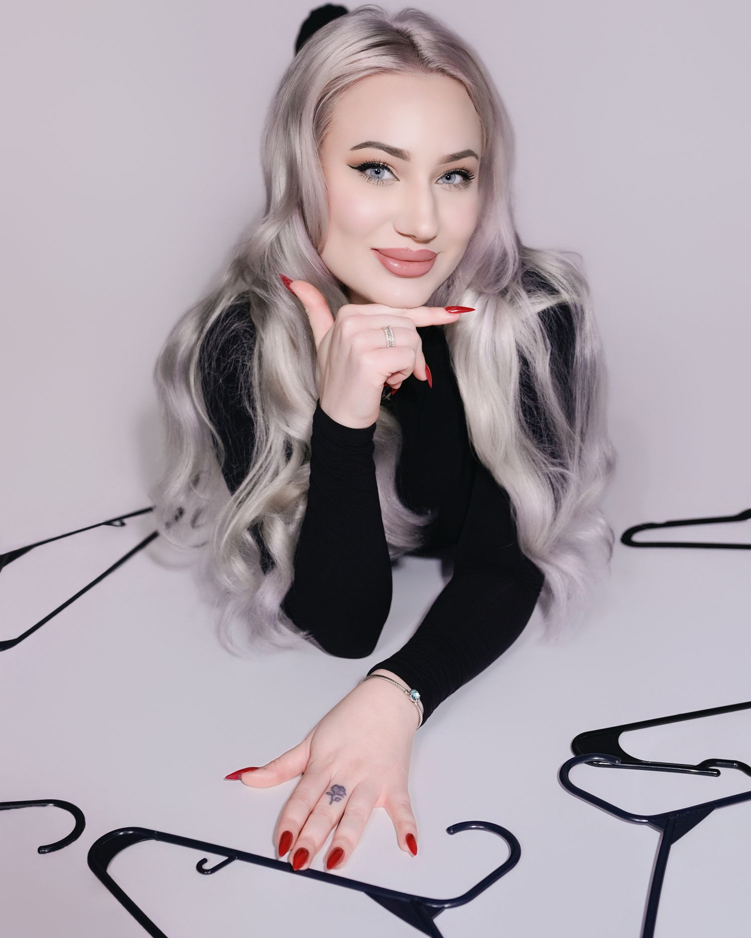 Portrait of someone with long wavy silver hair wearing black, posing against light background with red nail polish.