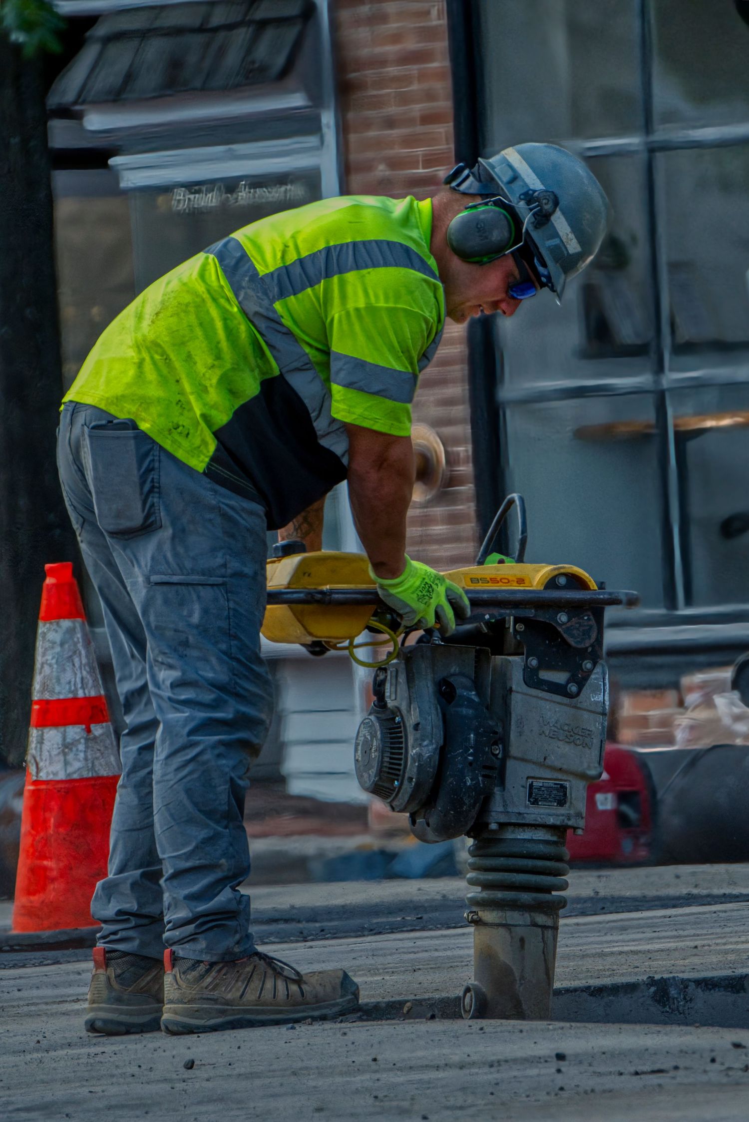 Construction worker in safety vest operates a gas-powered compactor on urban street with traffic cone nearby.