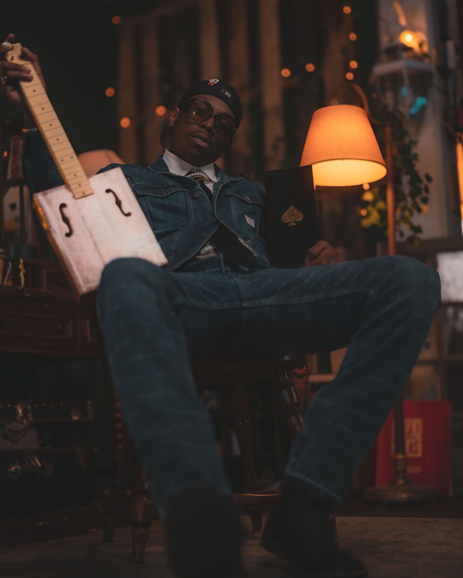 A musician in dark denim relaxes in a dimly lit room with a custom cigar box guitar and warm lamp glow.