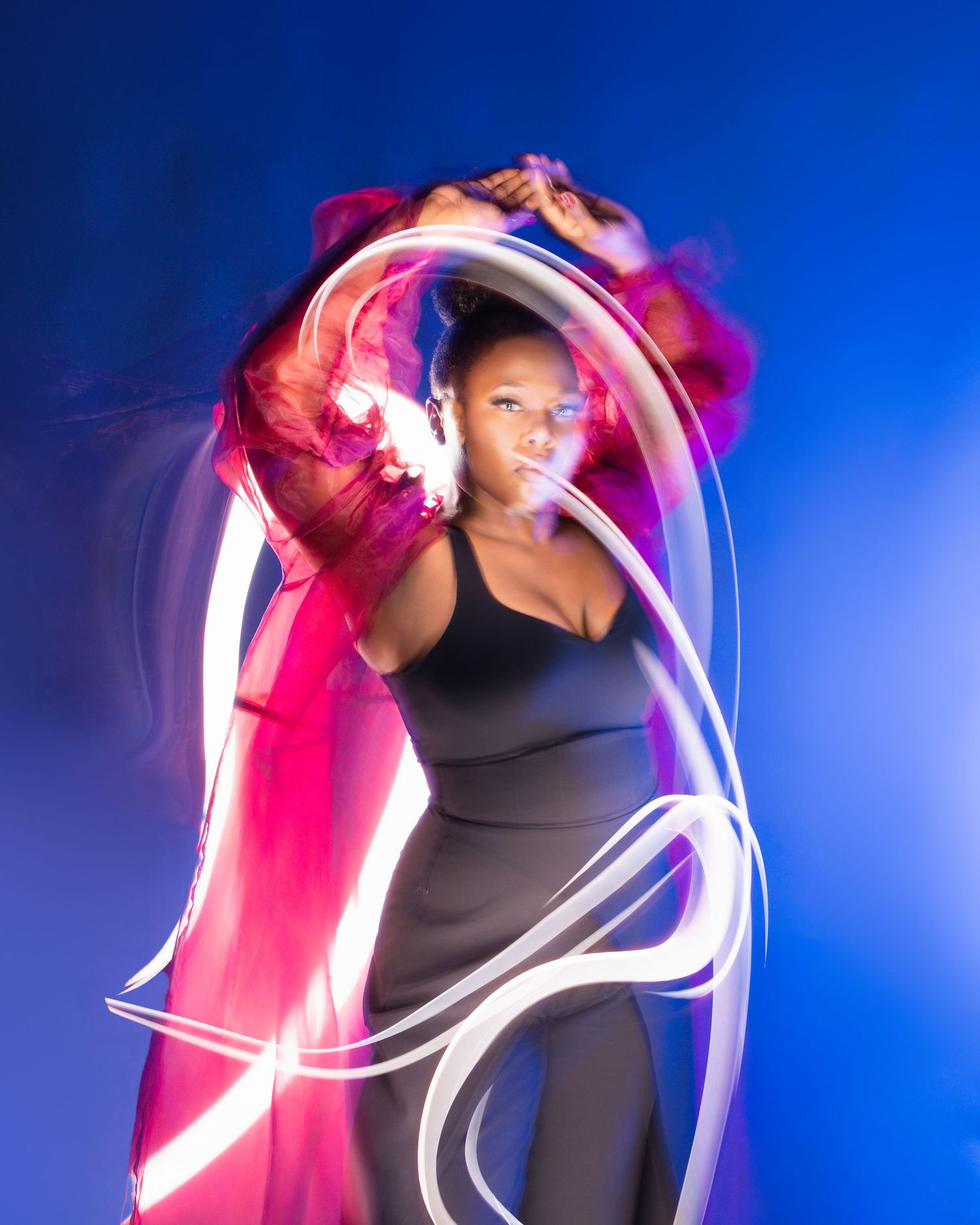 Dynamic dancer in black dress creates light trails with glowing white ribbons against vibrant blue backdrop.